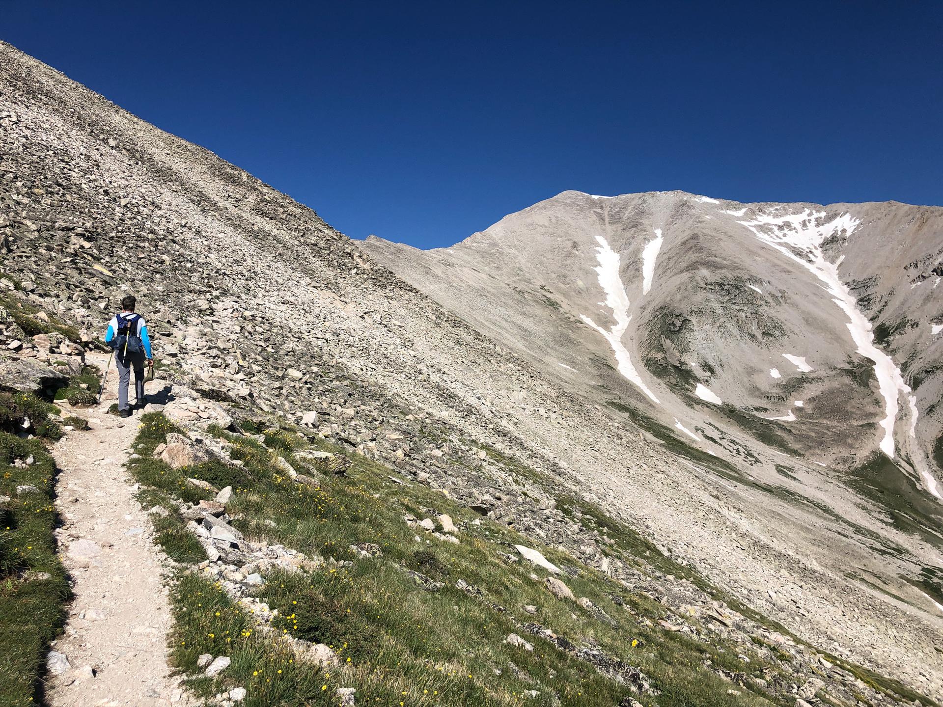 Hiker on a dirt trail beside a steep, rocky mountain slope on a sunny day.
