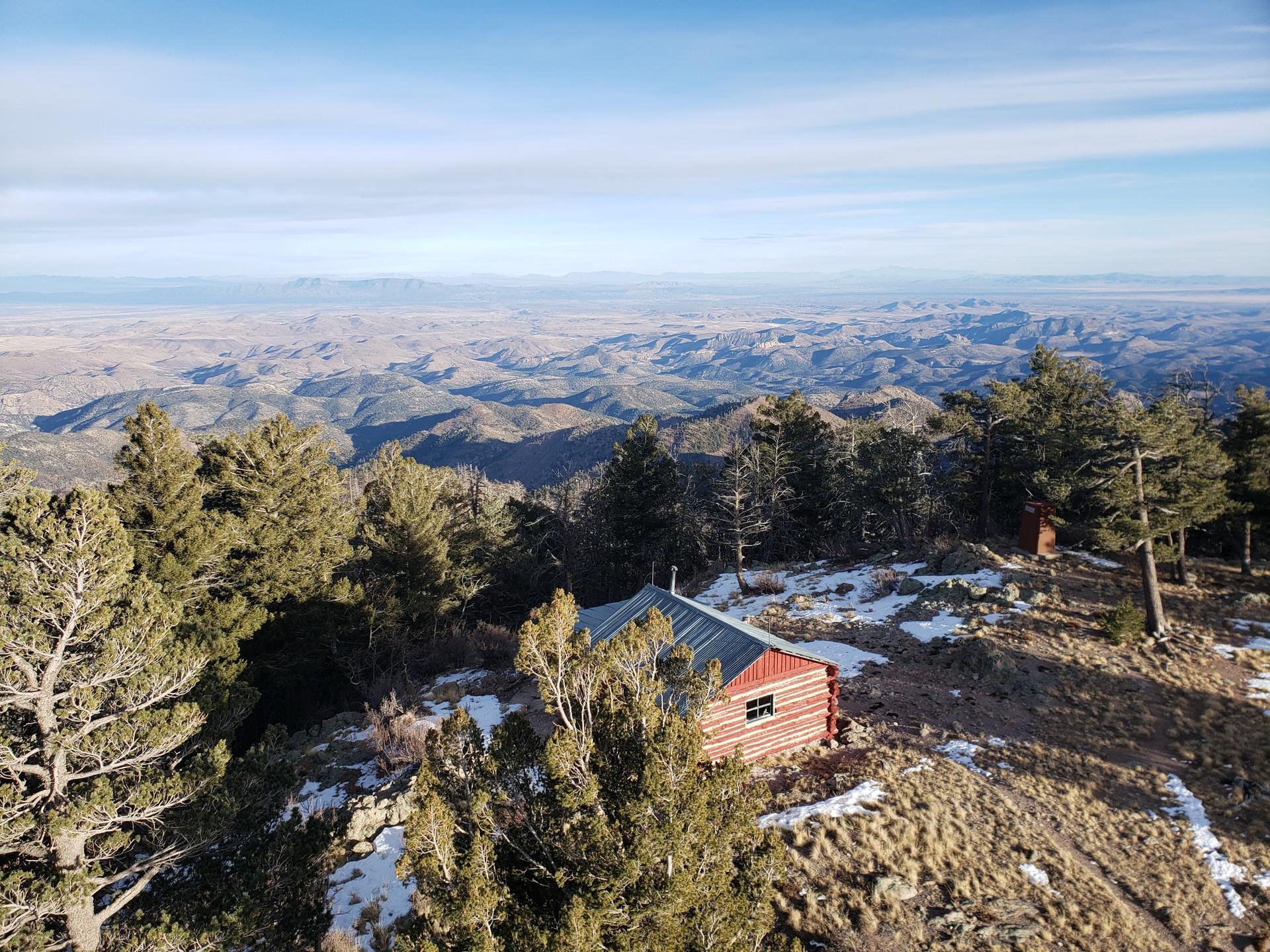 A red cabin nestled among pine trees overlooks a vast mountain landscape.