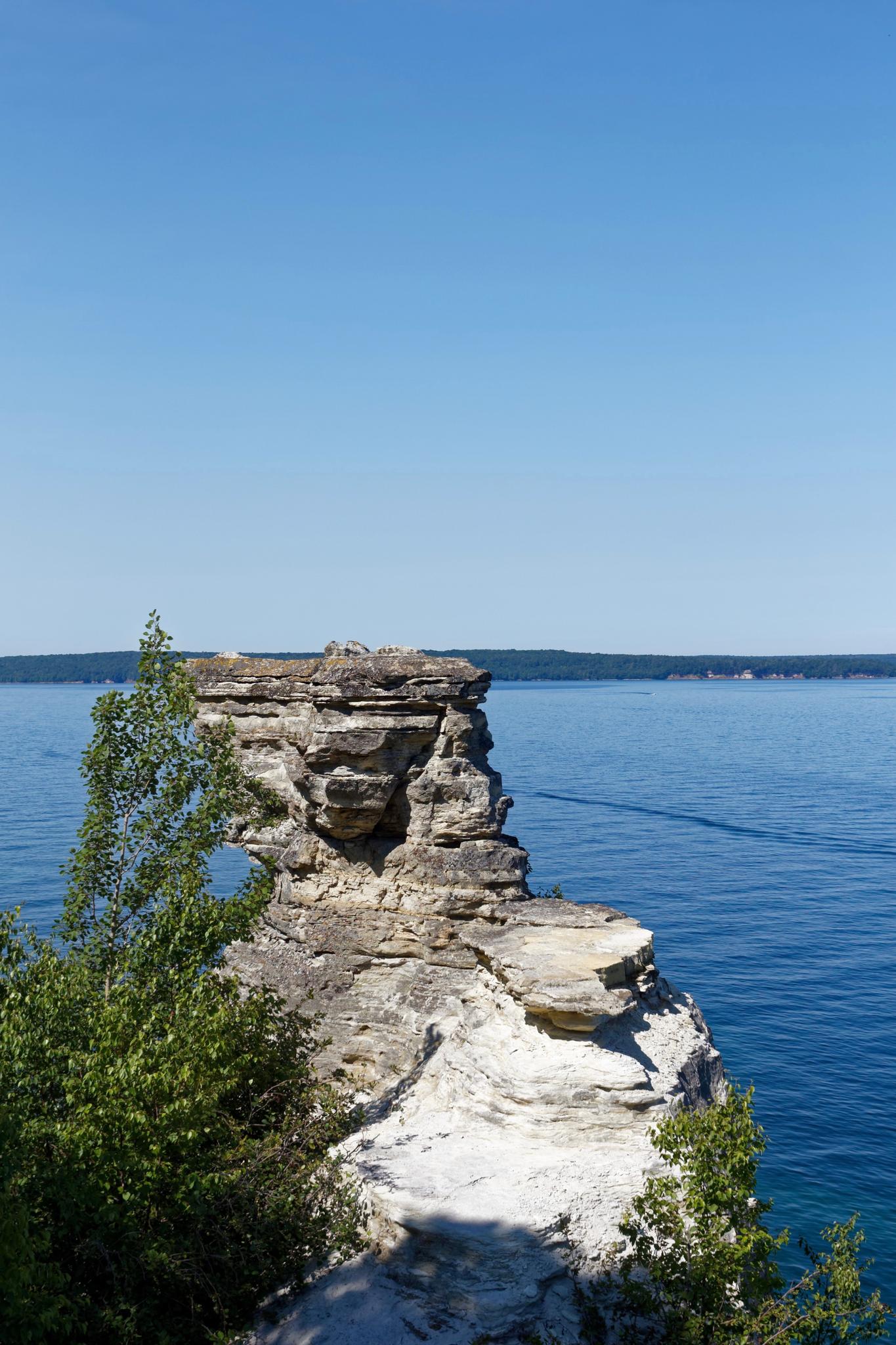 A tall, layered rock formation stands by calm blue water under a clear sky.