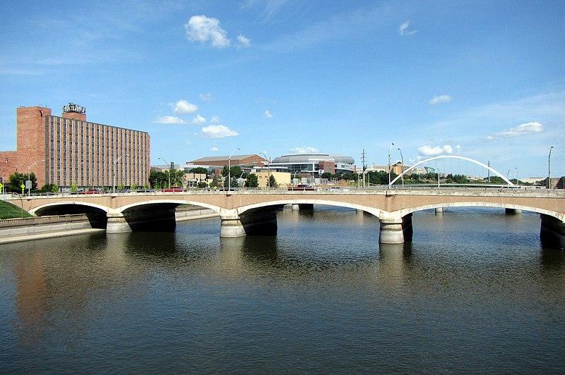 A multi-arched bridge spans a river, with city buildings visible under a bright sky.