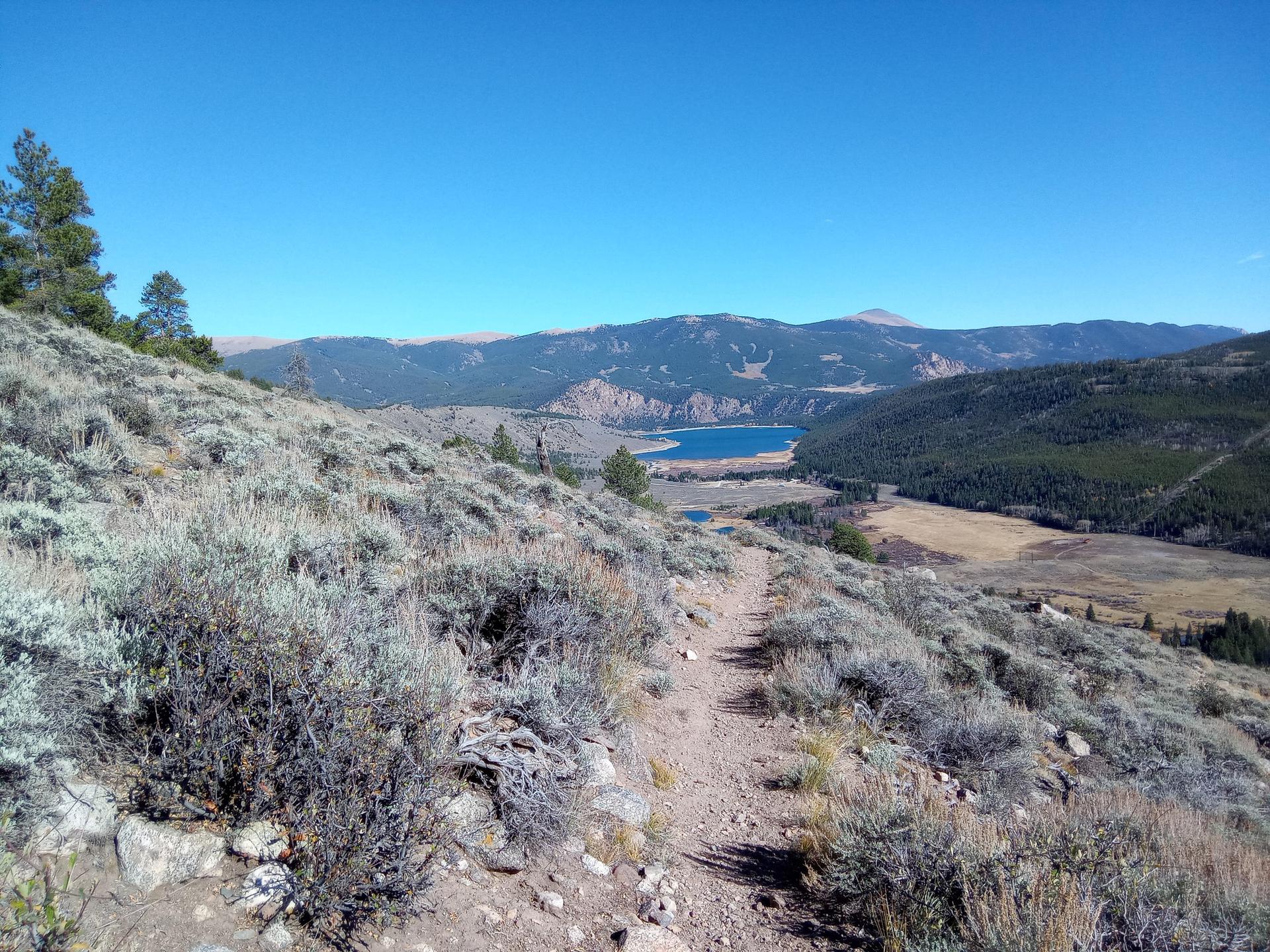 A dirt trail winds through a dry, brushy hillside overlooking a distant lake.