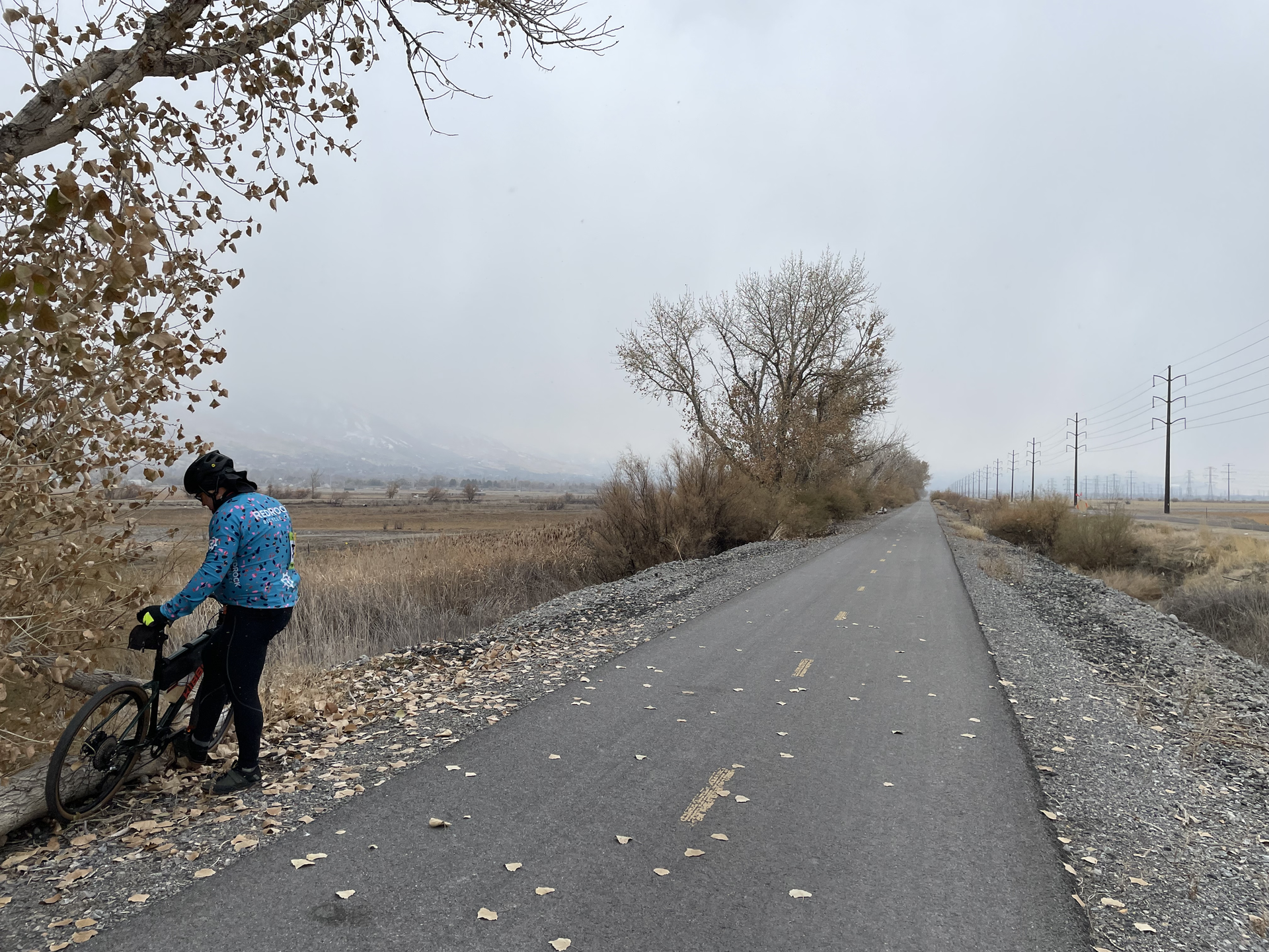 Cyclist stands beside a bike on a paved path on an overcast day.