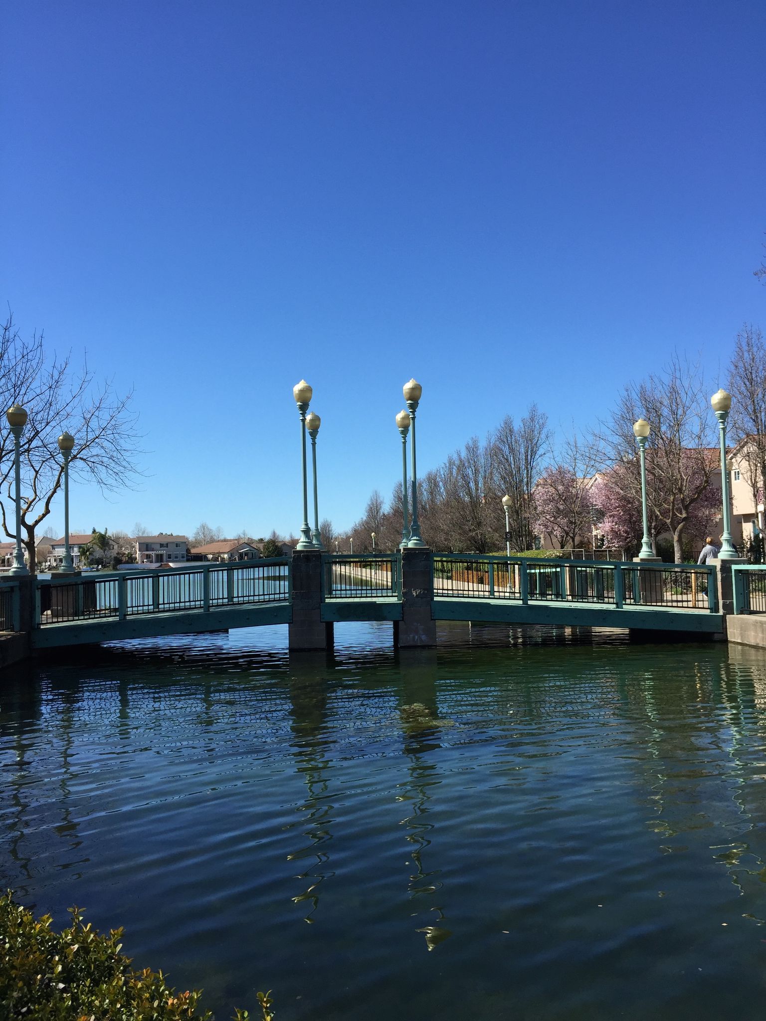 A green pedestrian bridge spans a canal on a bright day, with a person walking. - Bartholomew Park Loop