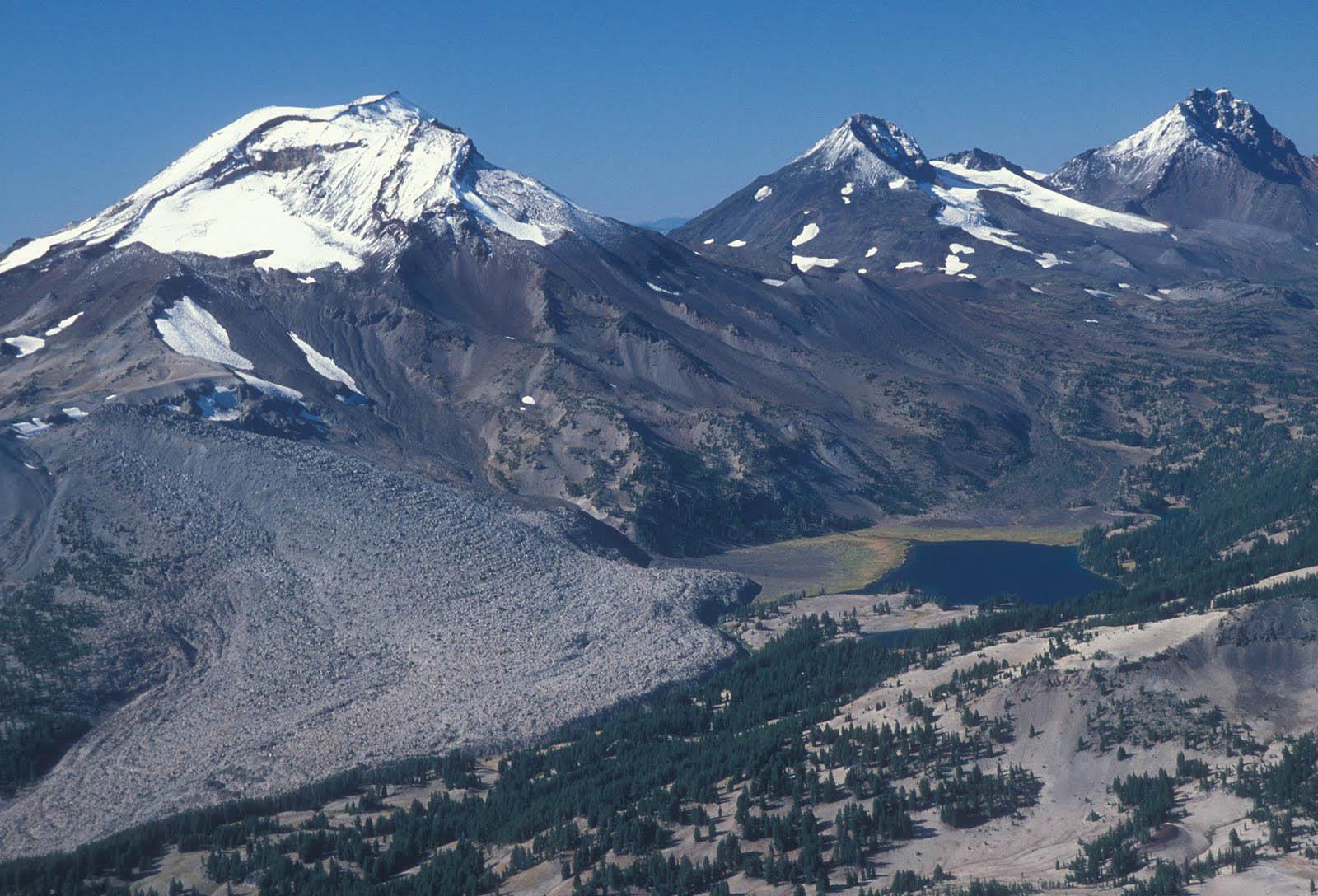 Three majestic snow-capped mountains overlook a rocky, forested valley with a small lake.