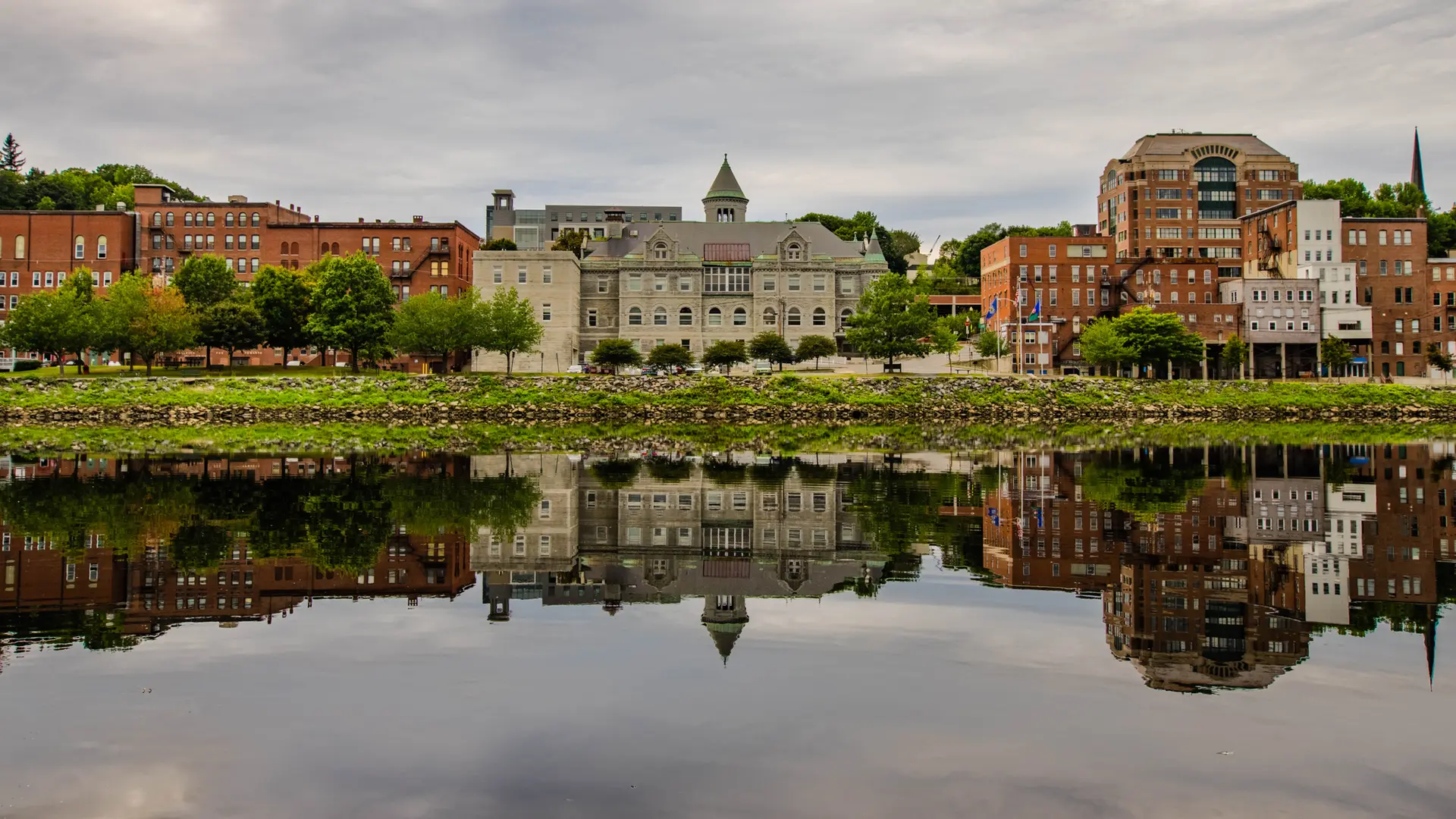 Riverside city buildings and trees perfectly reflected in calm water under a gray sky.