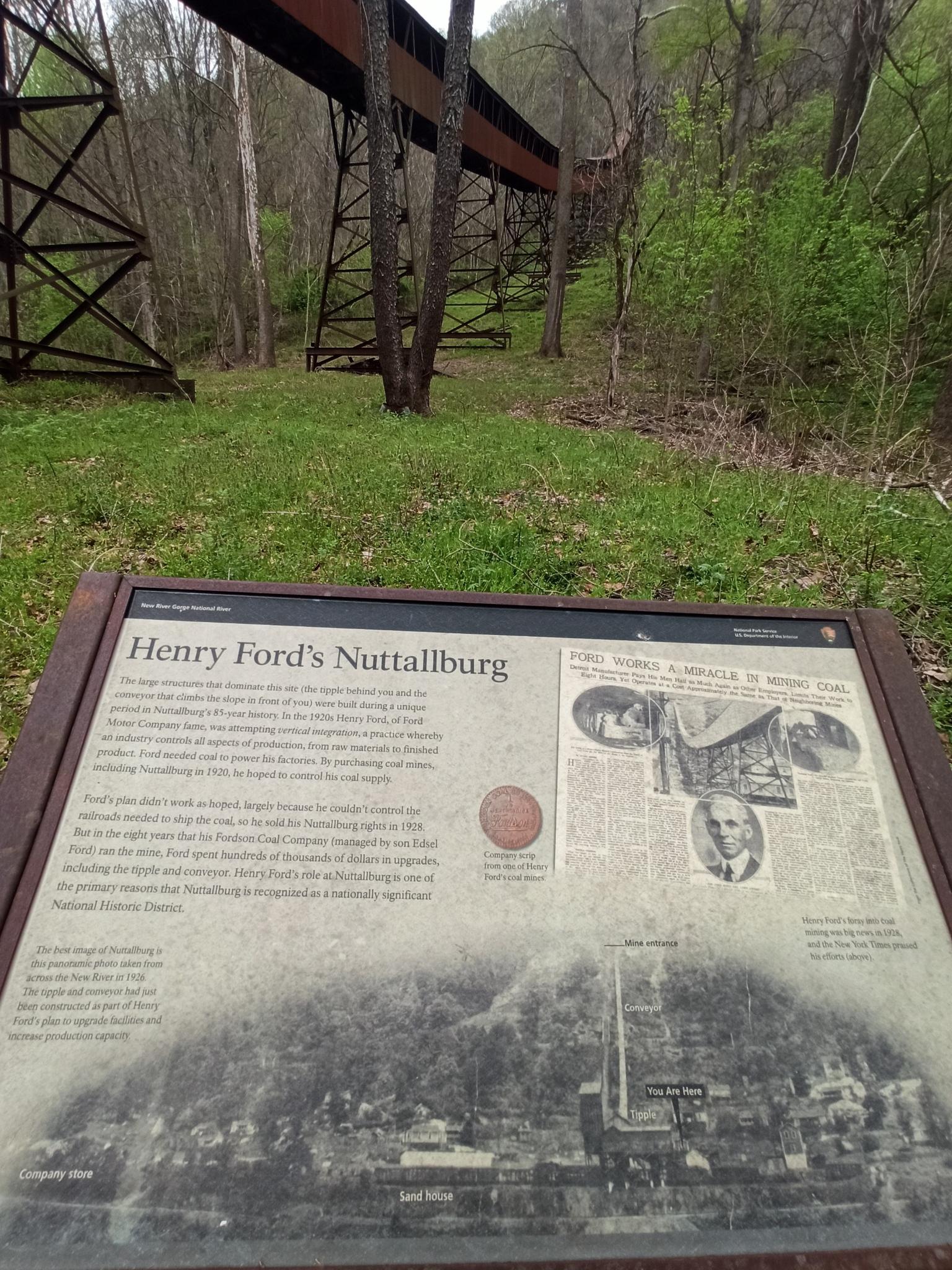 An informational sign stands before a large metal conveyor structure in a forest.