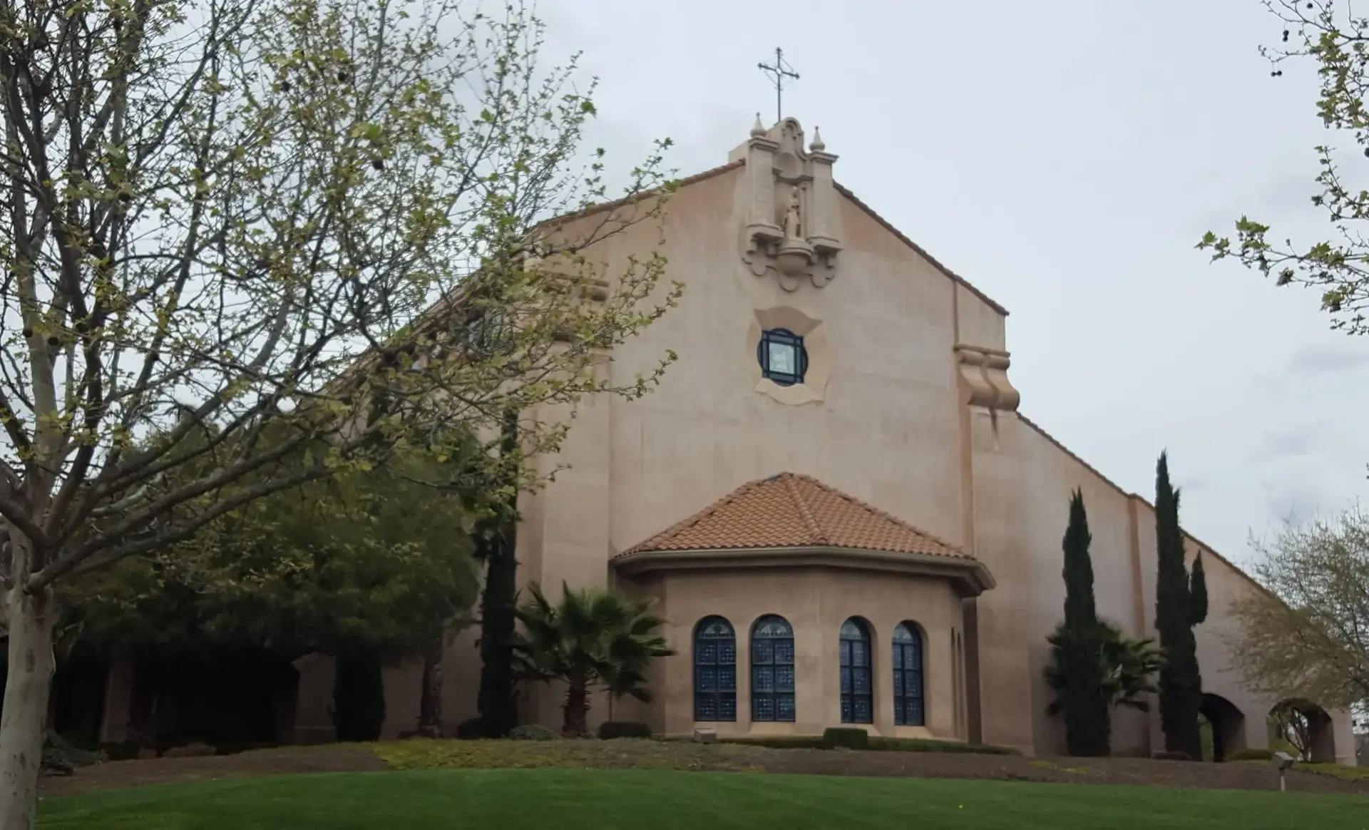 Large mission-style building with cross, green lawn, and trees under an overcast sky.