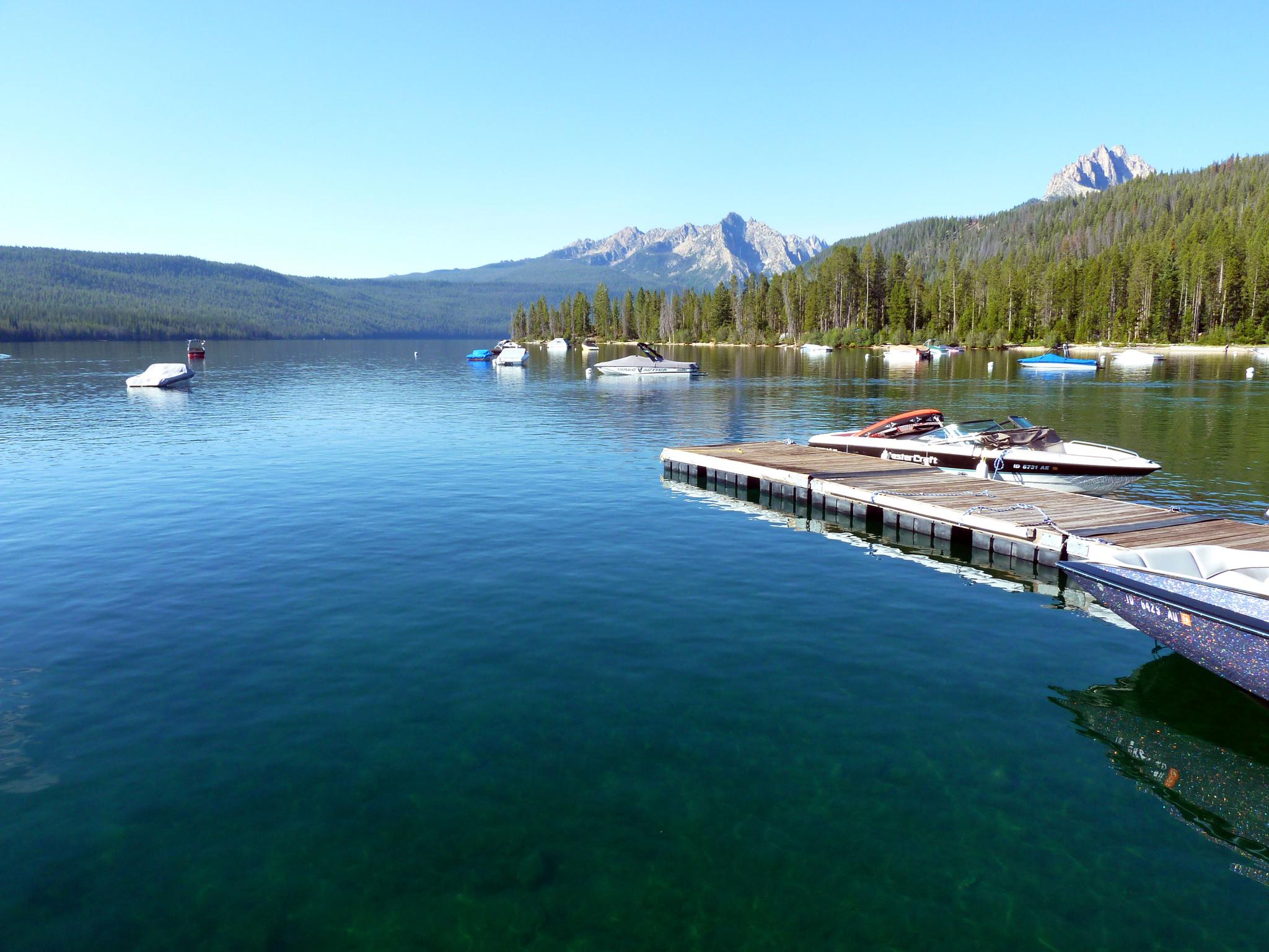 Boats on a clear blue lake with forested mountains under a sunny sky.
