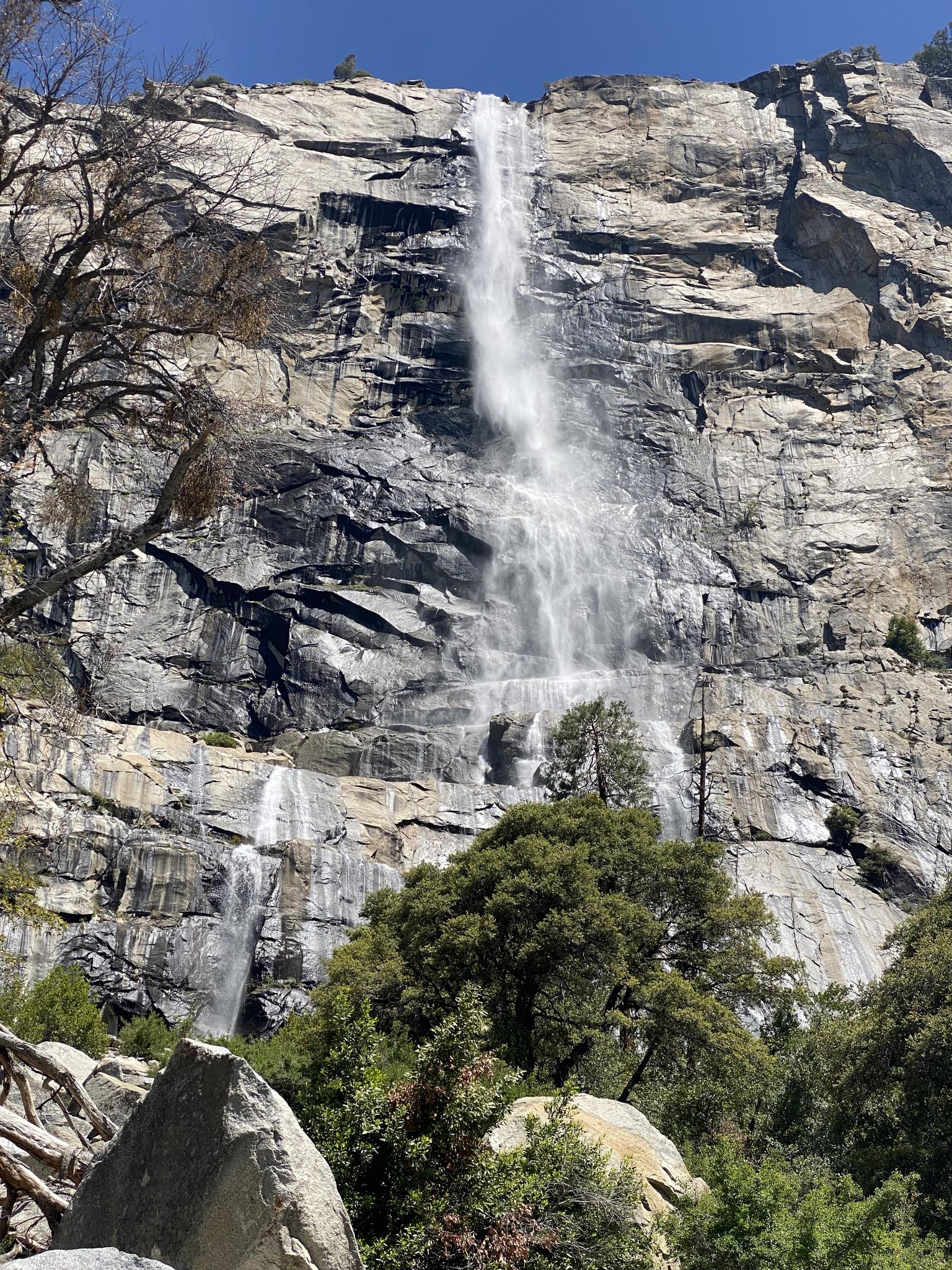 Tall waterfall cascades down a massive grey cliff, surrounded by green trees.