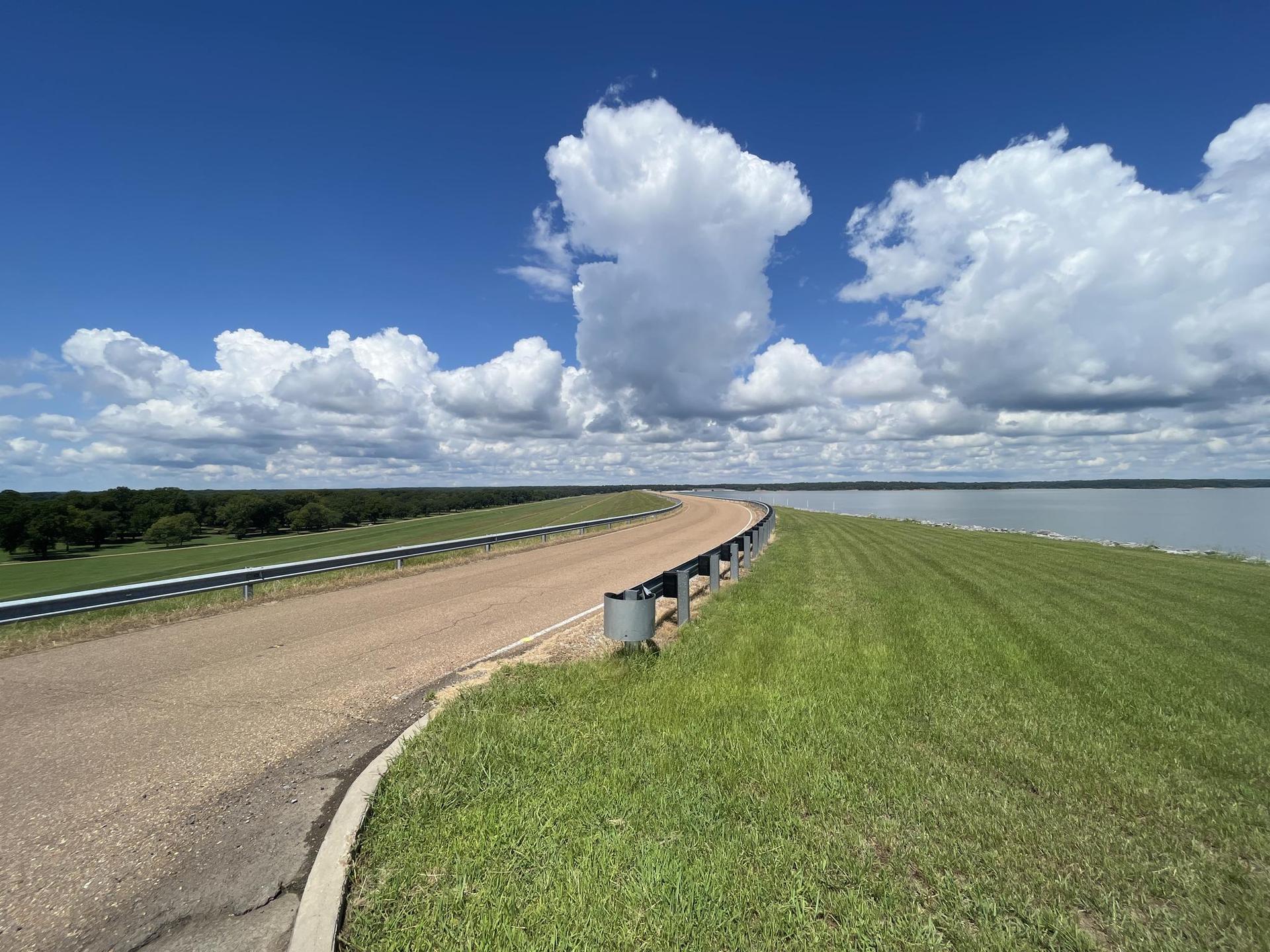 Curved road on a grassy embankment next to water under a bright, cloudy sky.