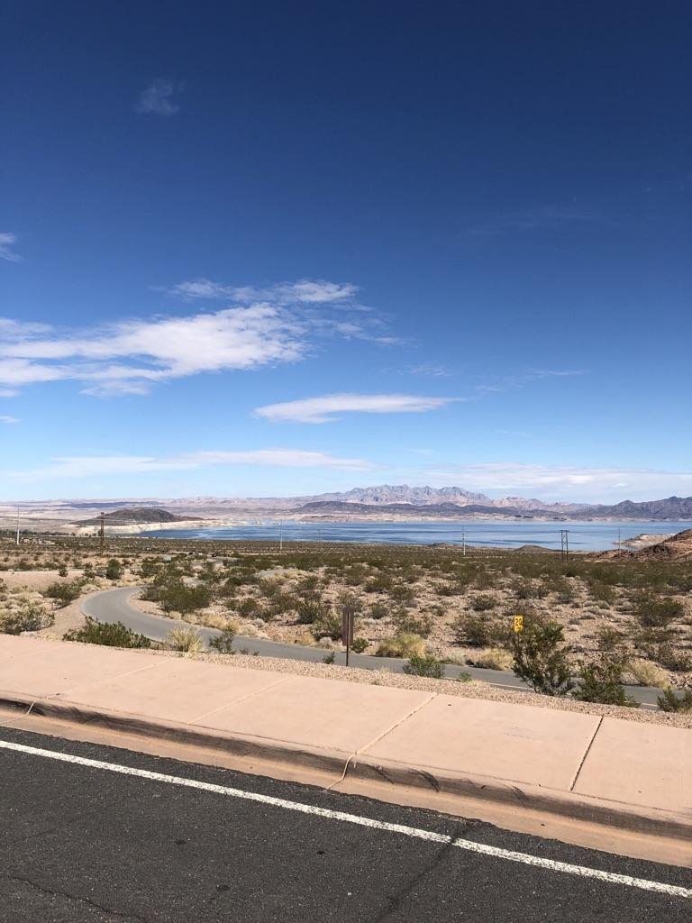 Desert landscape with a curving road, large lake, and distant mountains under blue sky.