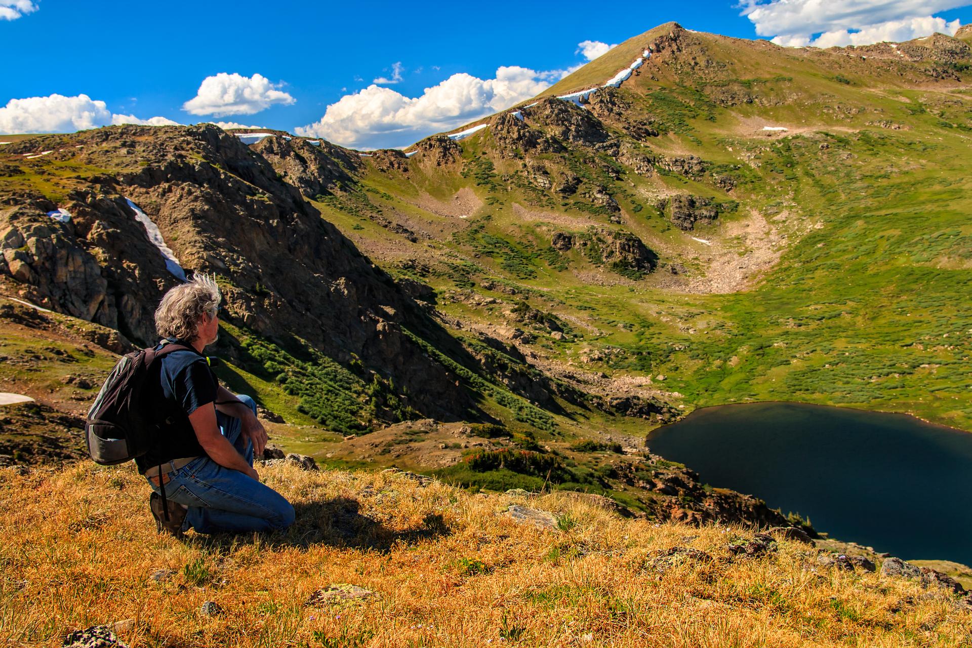 Hiker sits on a golden hillside, admiring a mountain lake under blue sky.