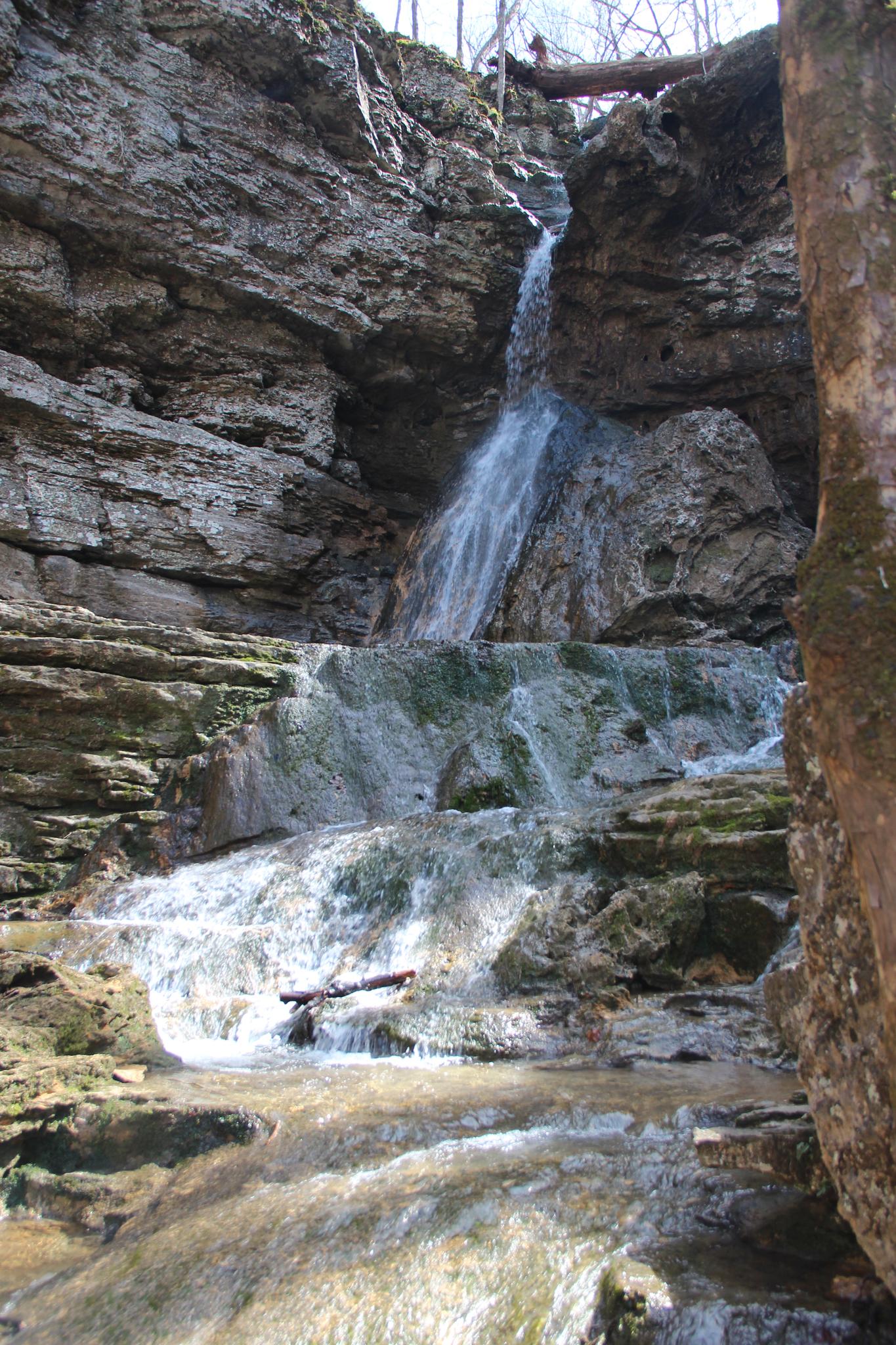 A multi-tiered waterfall cascades down rocky cliffs under bright daylight.