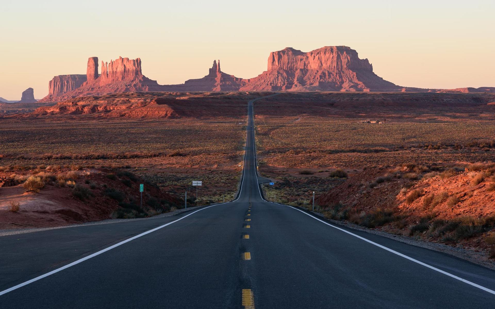 A long road leads to distant red rock formations at sunset.