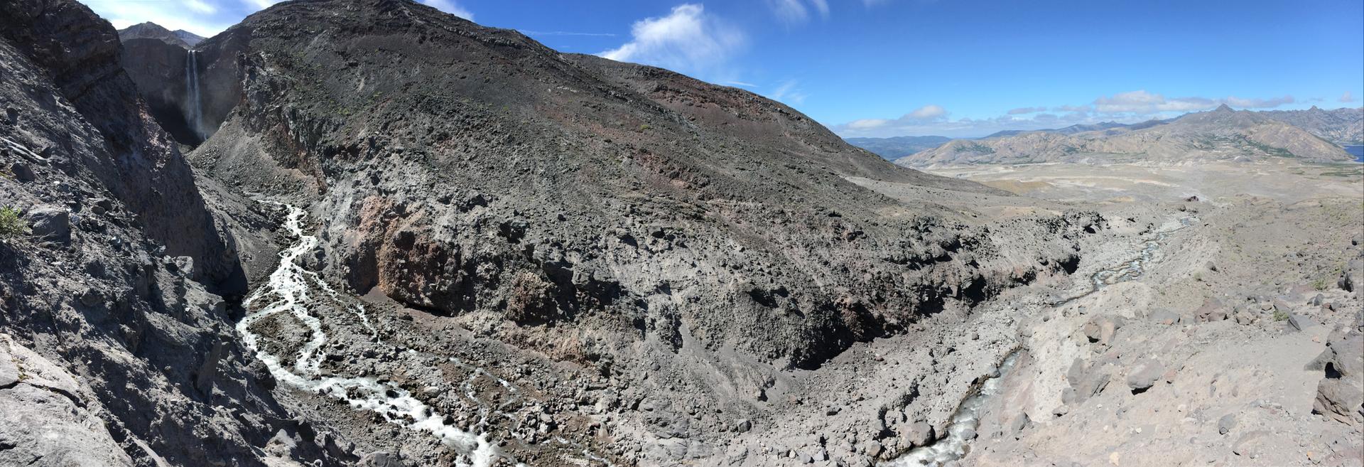 Rocky mountain landscape with a winding river and distant waterfalls under blue sky.