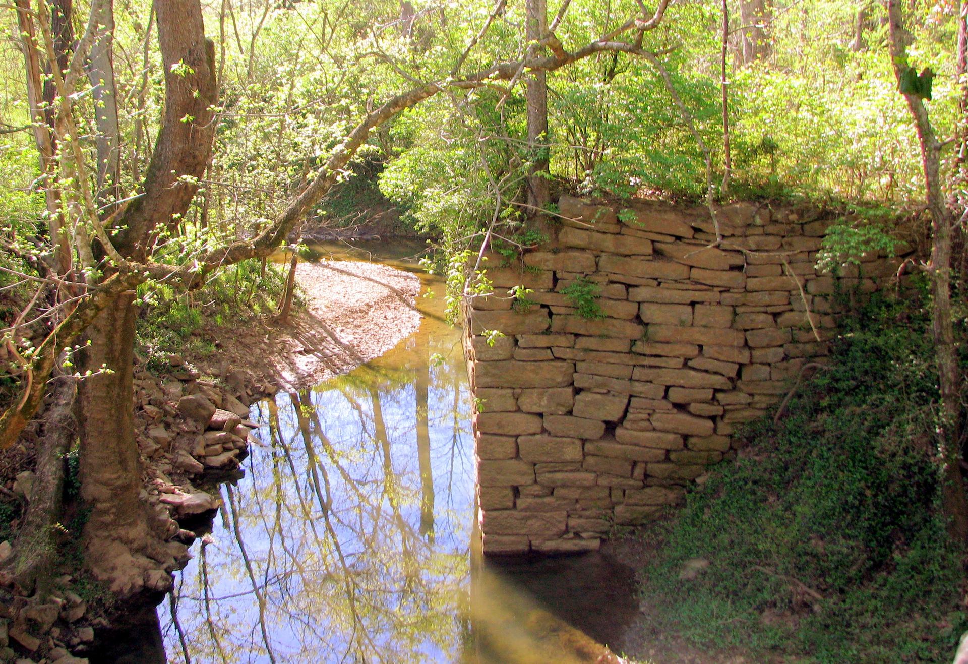 A sunlit stream flows past an old stone wall in a green forest.