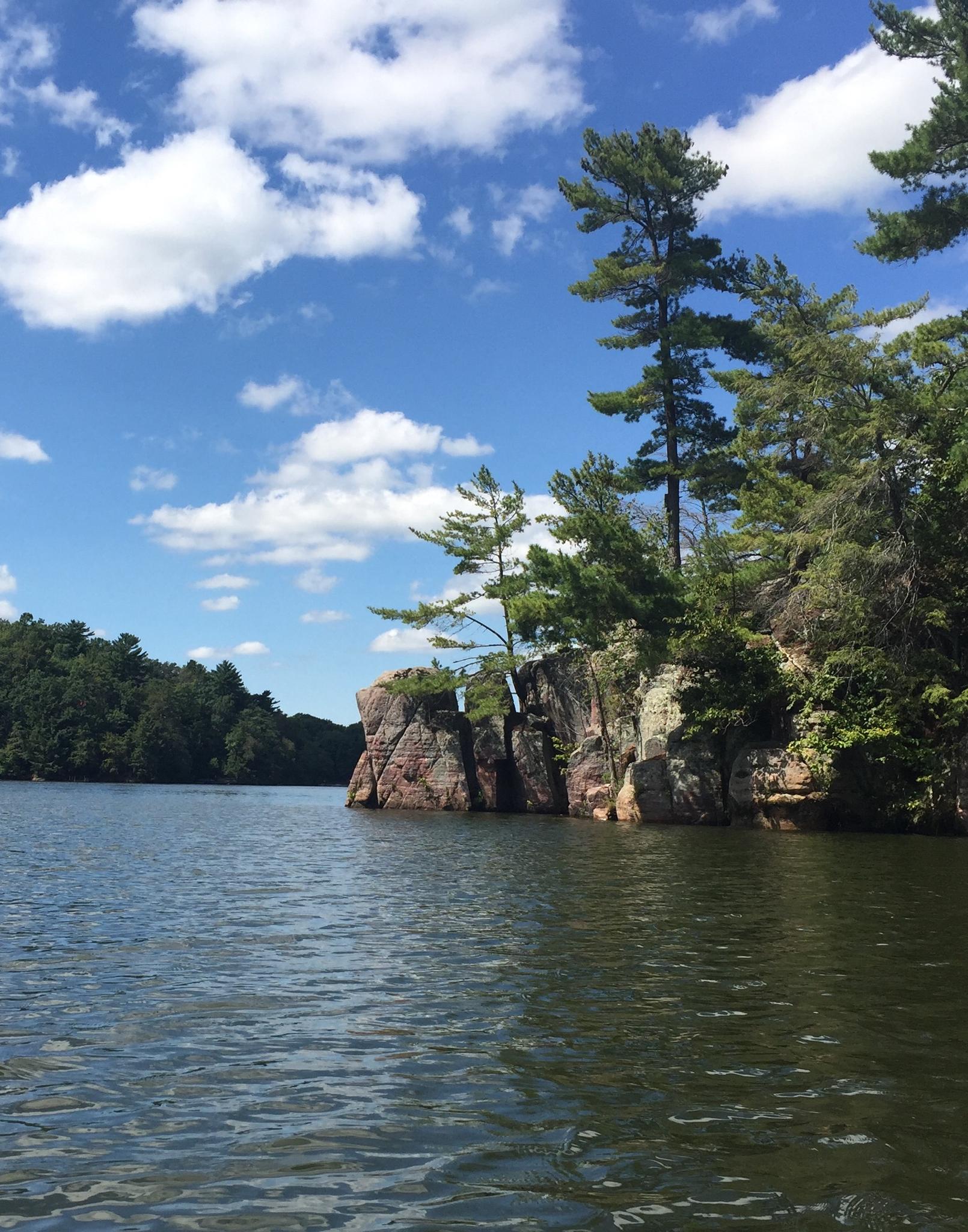 Blue water, rocky cliffs with pine trees, and a bright, cloudy sky.