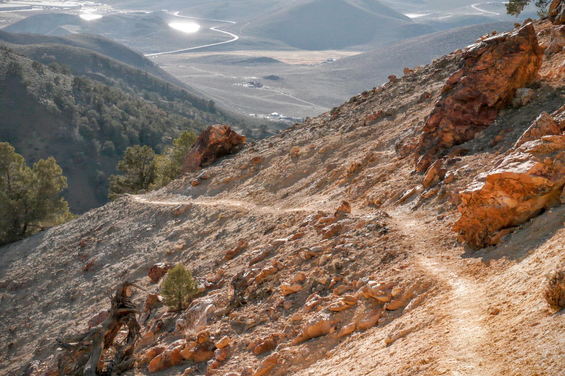 A narrow dirt trail traverses a steep, rocky mountainside overlooking a valley.