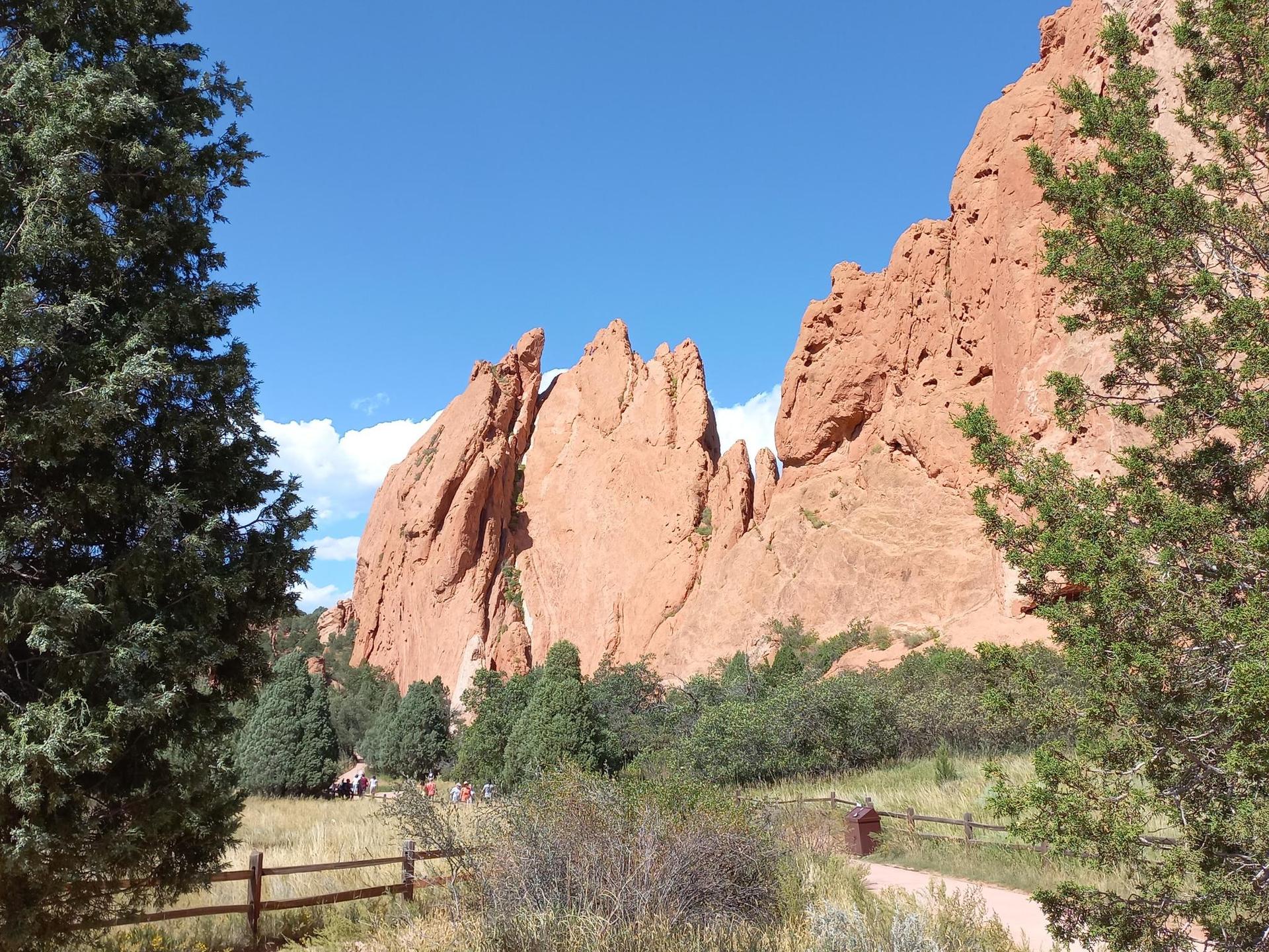 Red rock formations frame a path and distant people through green trees under a blue sky.