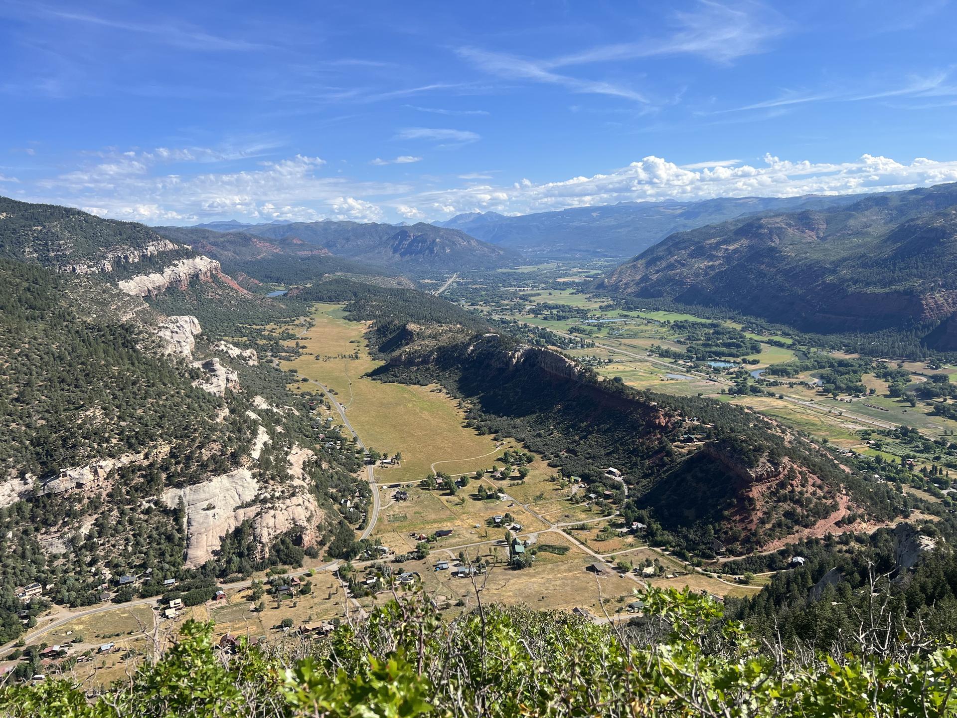 Bright daylight illuminates a wide green valley with settlements, surrounded by mountains.