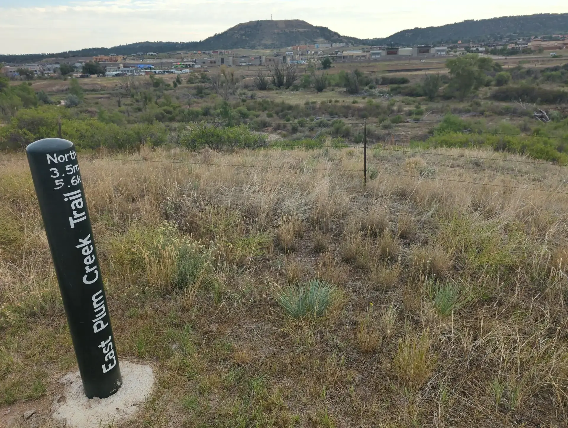 A black trail marker in a dry, grassy field with distant hills under an overcast sky.