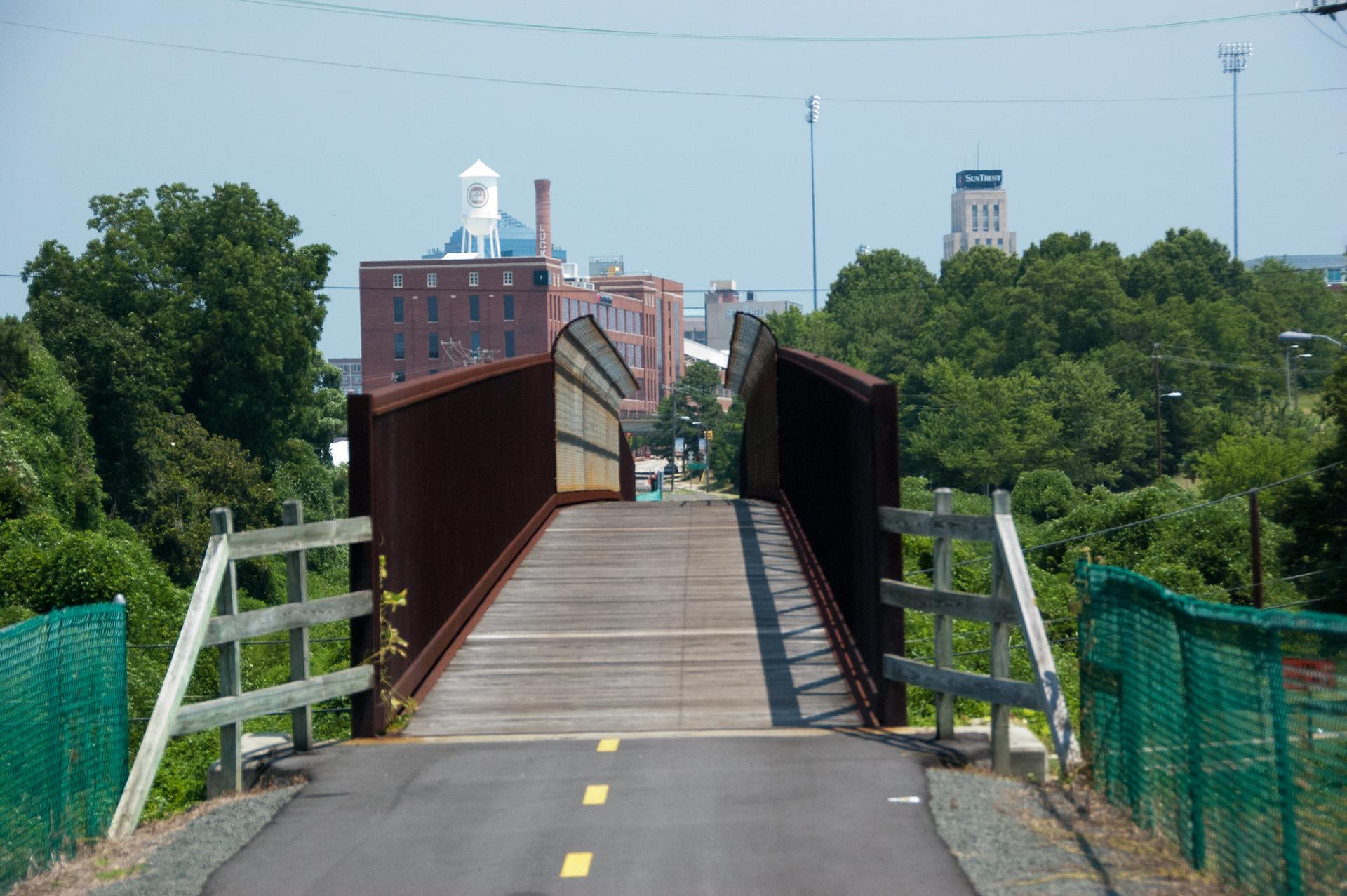 Paved path leads to a wooden pedestrian bridge under a bright, sunny sky.