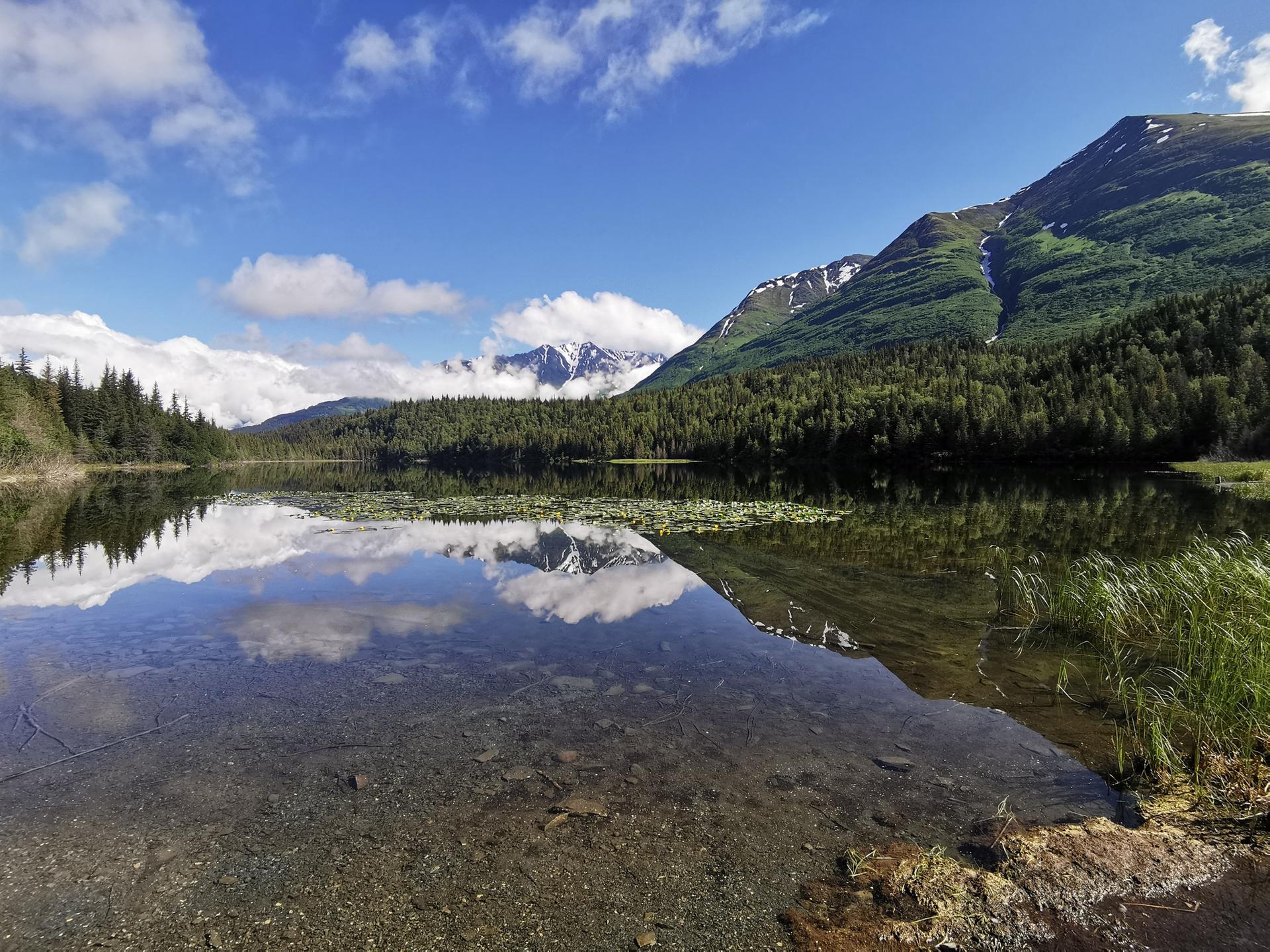 Sunny day over a clear lake reflecting mountains and clouds.
