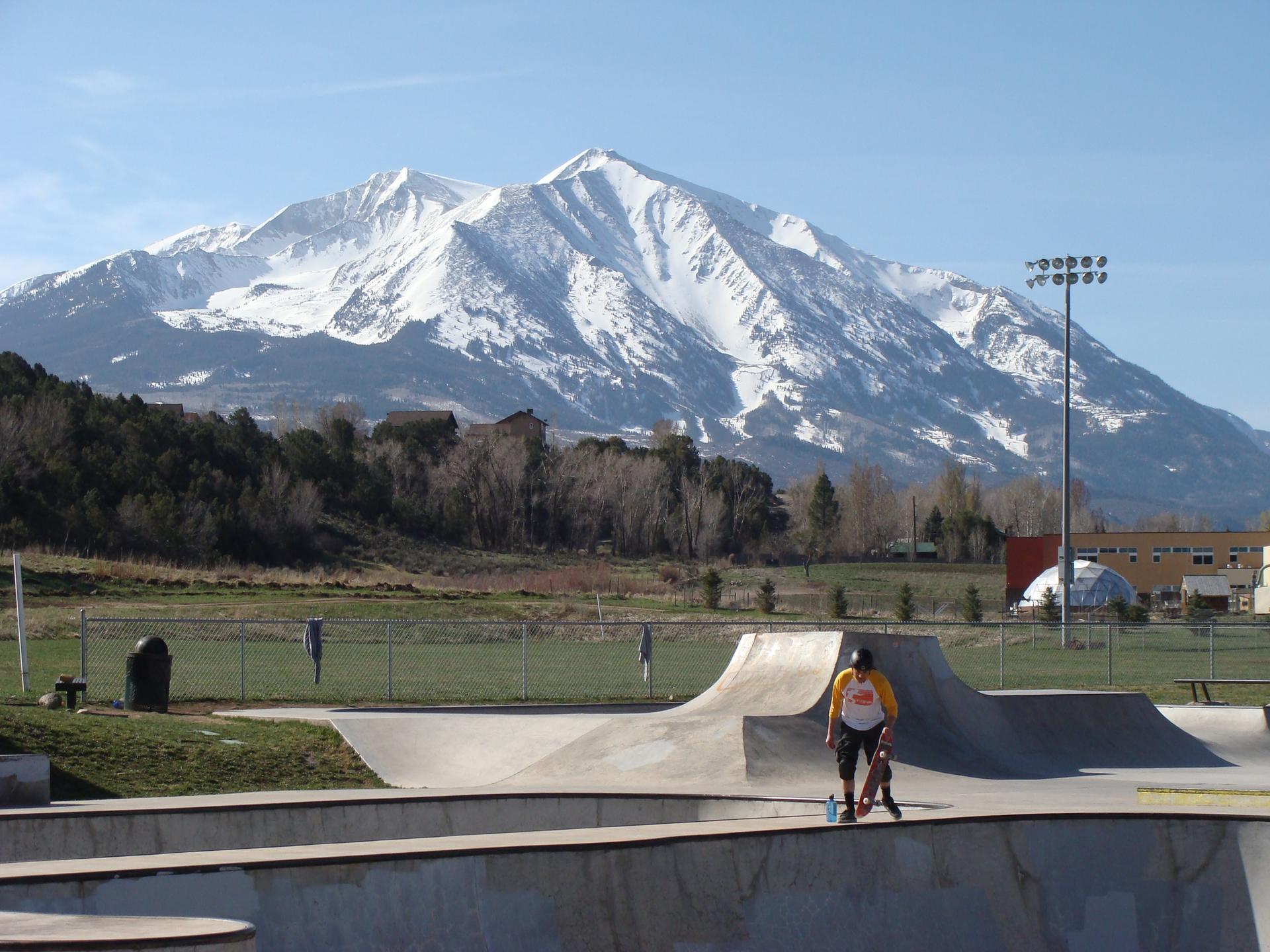 Person skateboarding in a sunny skatepark with a large snow-capped mountain backdrop.