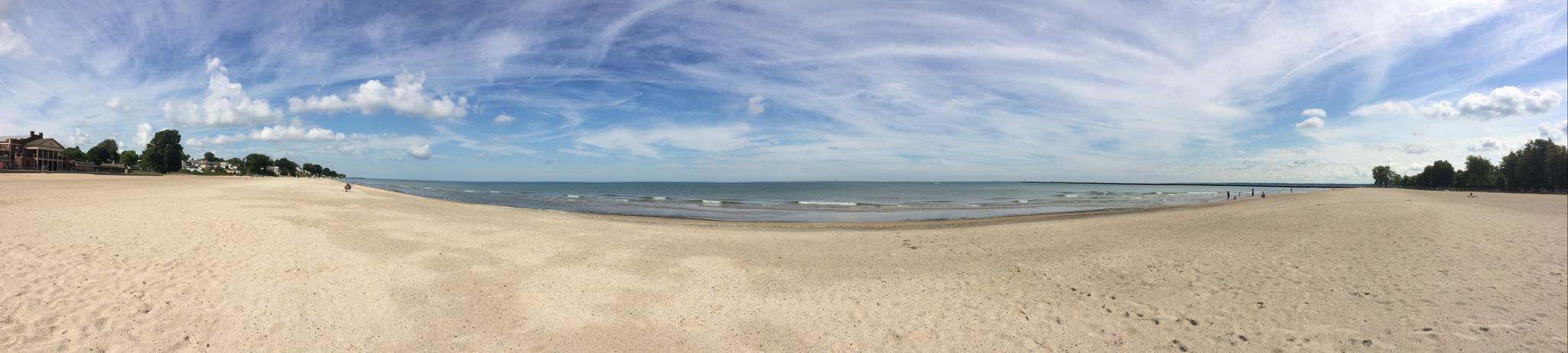 Panoramic view of a wide sandy beach, calm water, and a bright, cloudy sky.