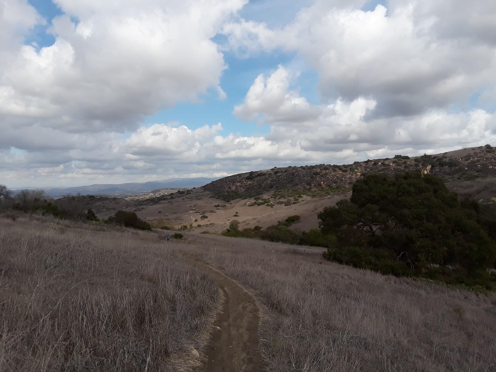 Winding dirt path through dry hills and brush under a cloudy sky.