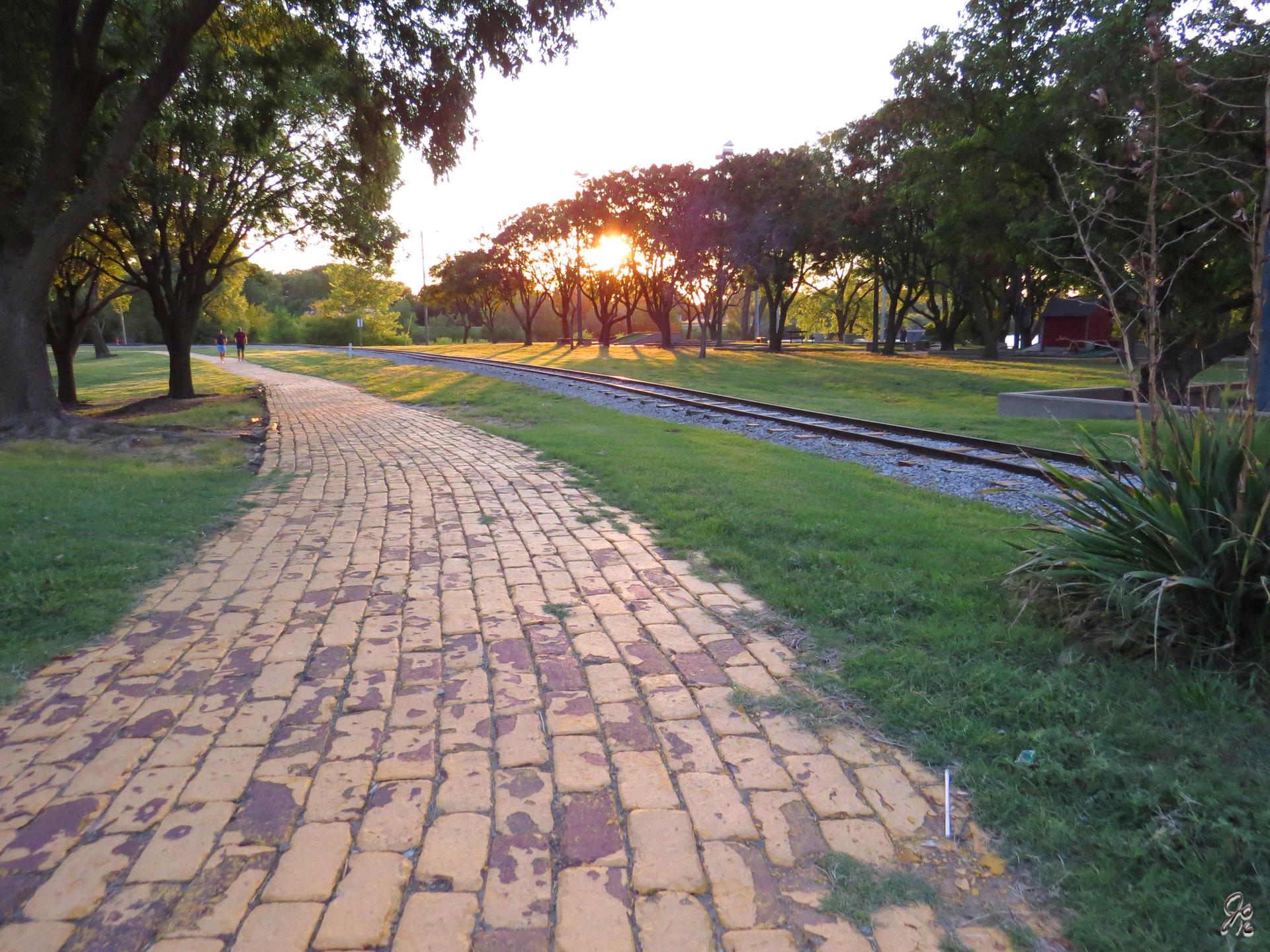 Golden hour light illuminates a brick path and railroad tracks through trees.