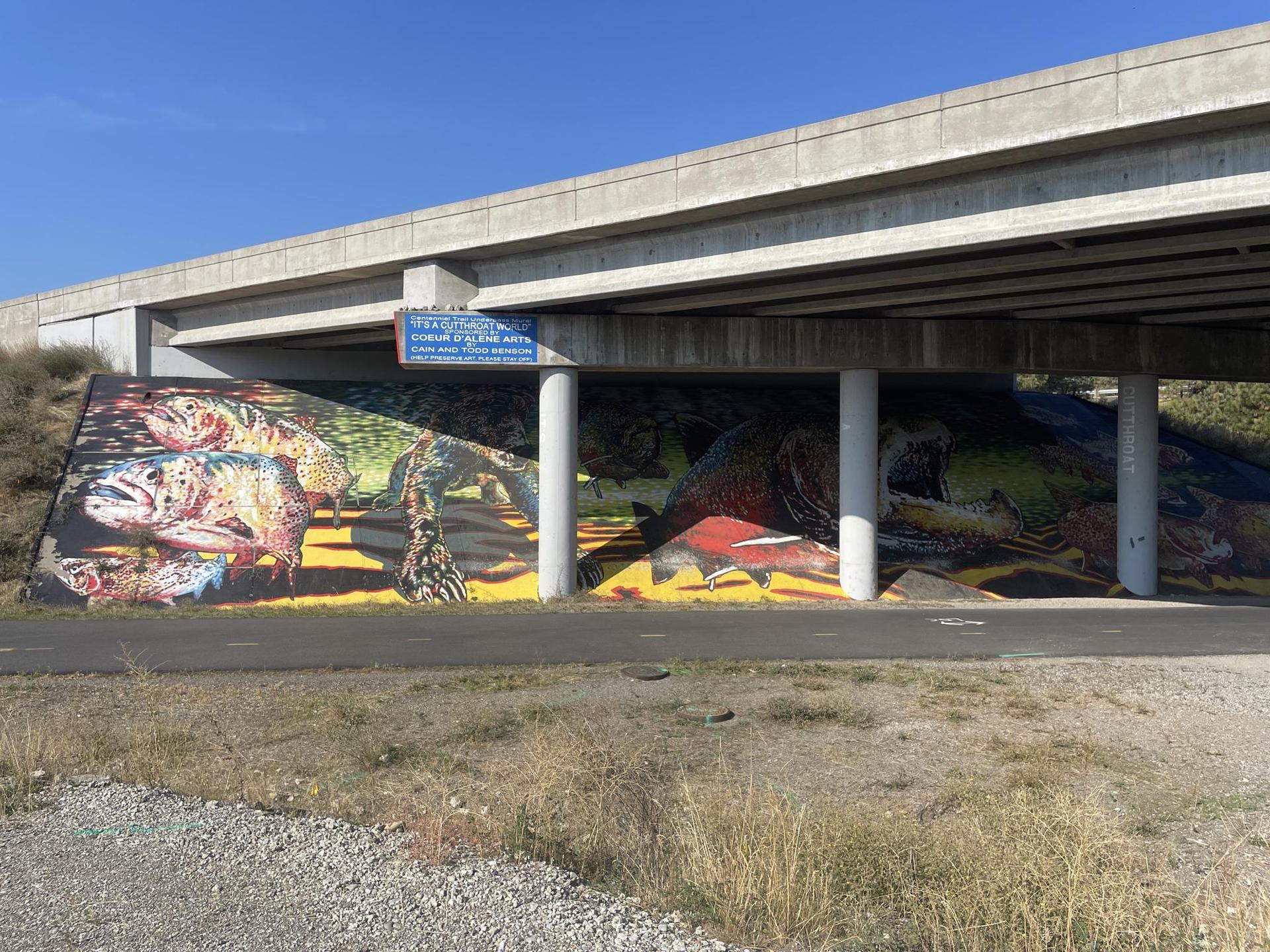 Colorful mural depicting nature under a concrete overpass on a sunny day.