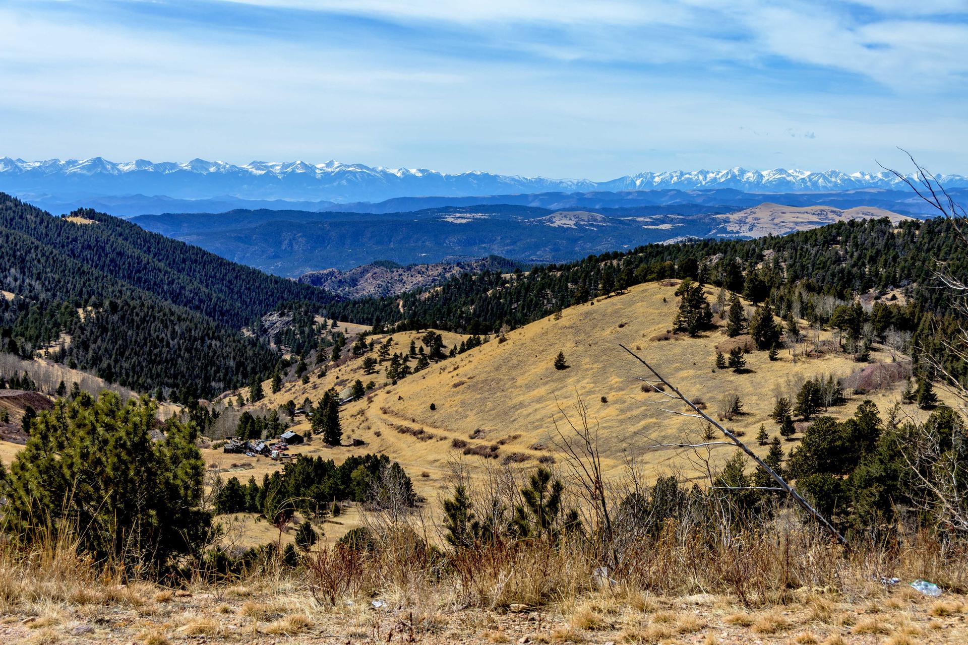 Vast mountain landscape with forested hills, dry slopes, and distant snow-capped peaks.
