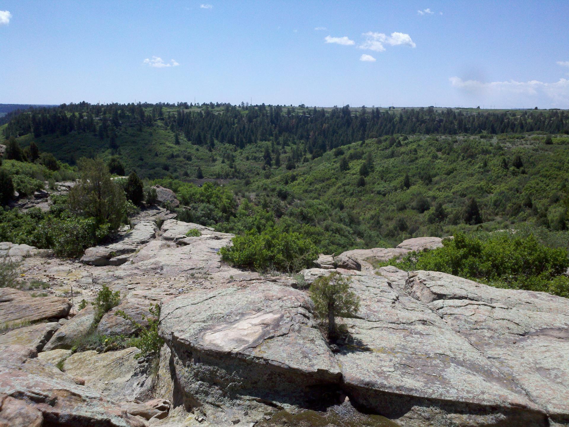 Rocky outcrop overlooks a lush green valley under a bright blue sky.