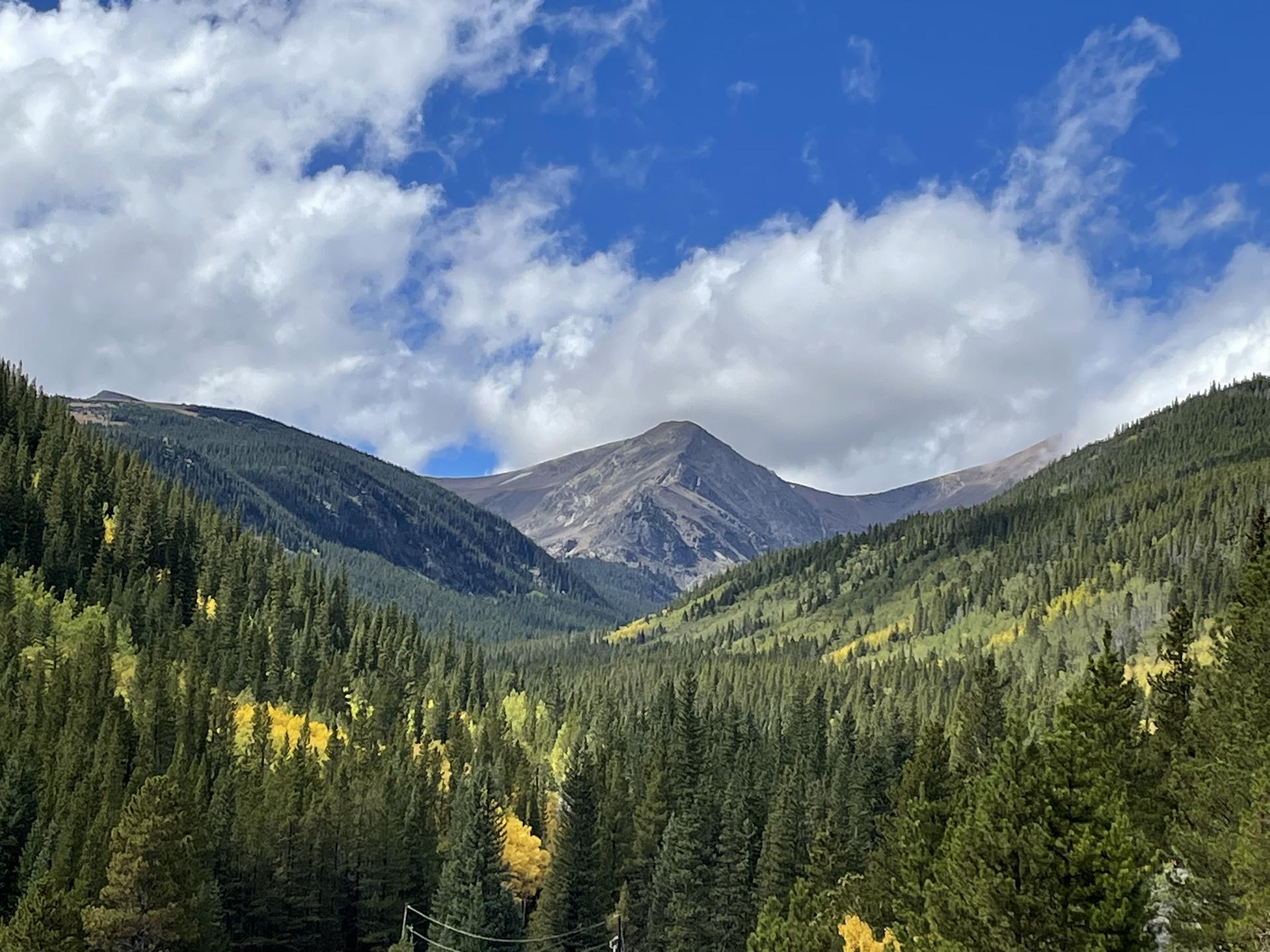 Forested mountain slopes with autumn colors under a bright, cloudy sky.