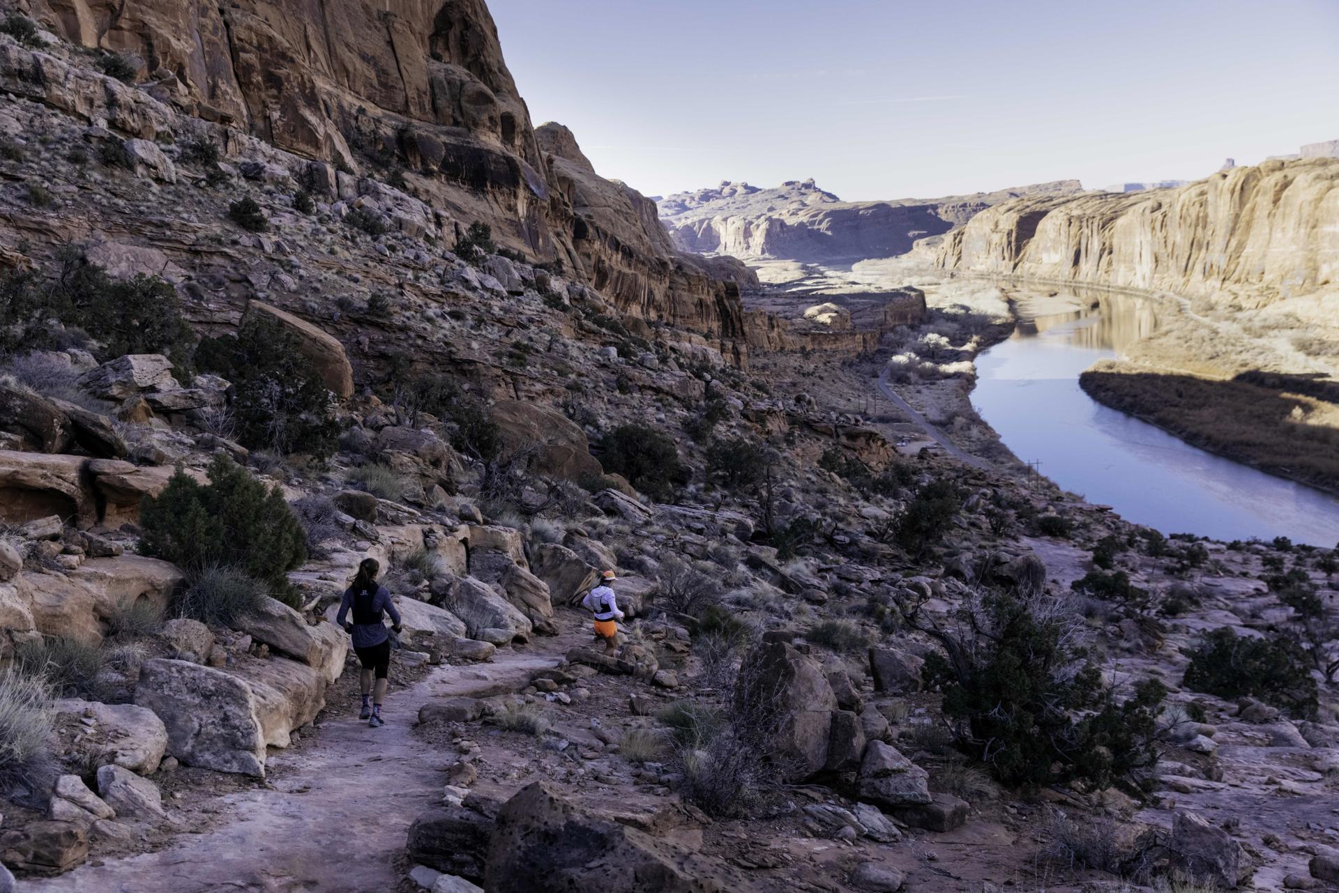 Two hikers on a rocky trail beside a river in a canyon.