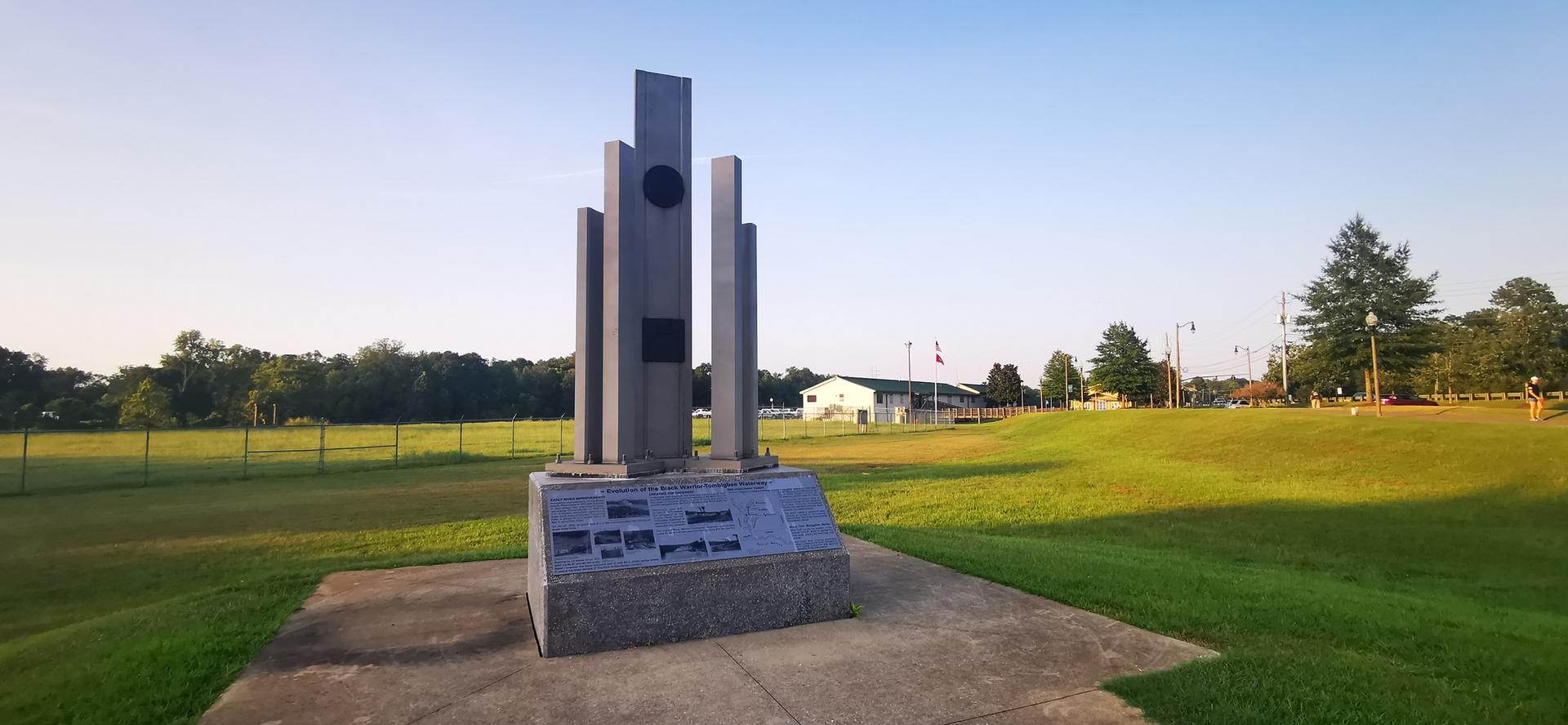 Tall stone monument in a sunny, expansive green field under clear sky.