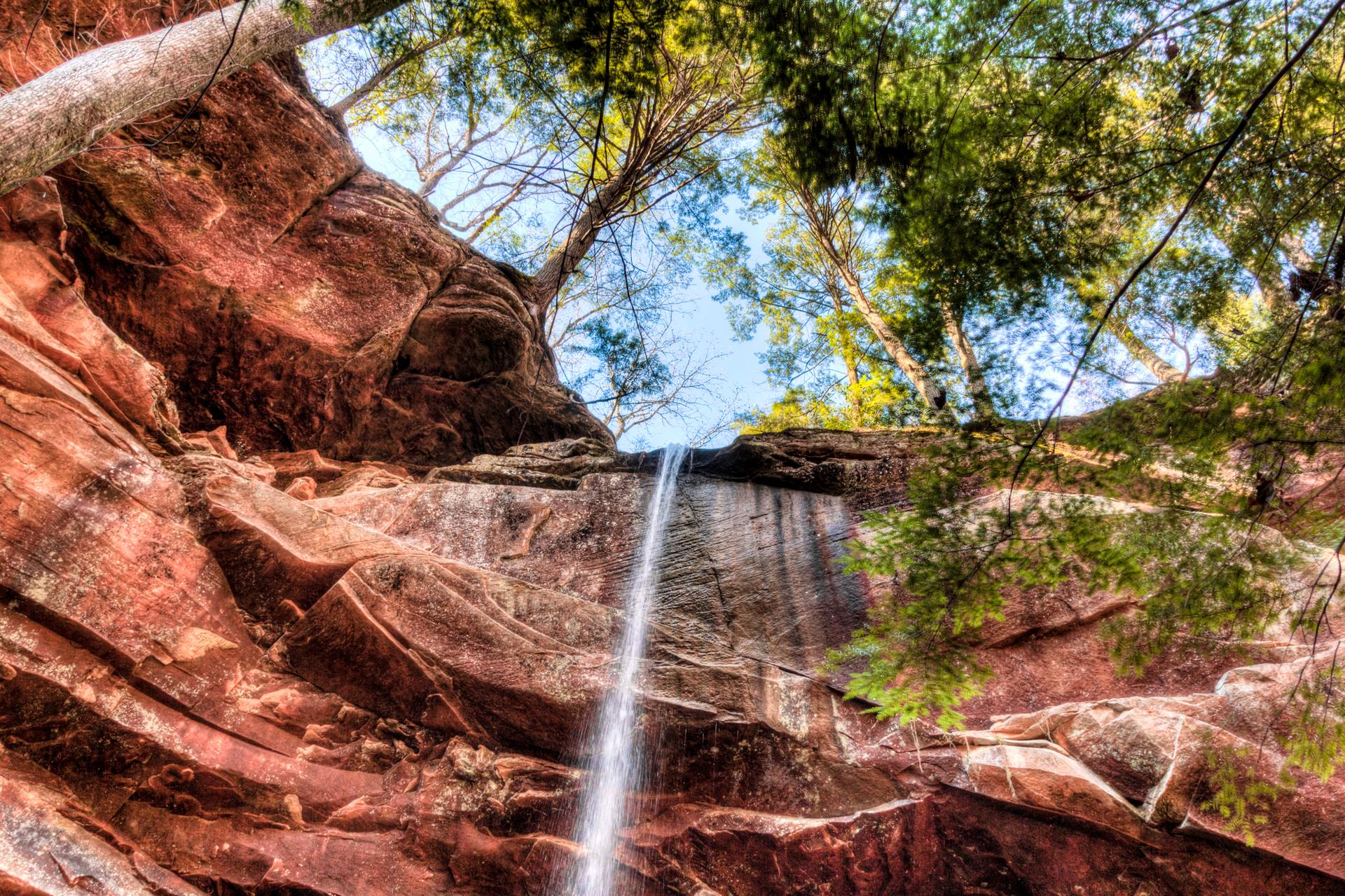 A narrow waterfall cascades down a red rock cliff under green trees.