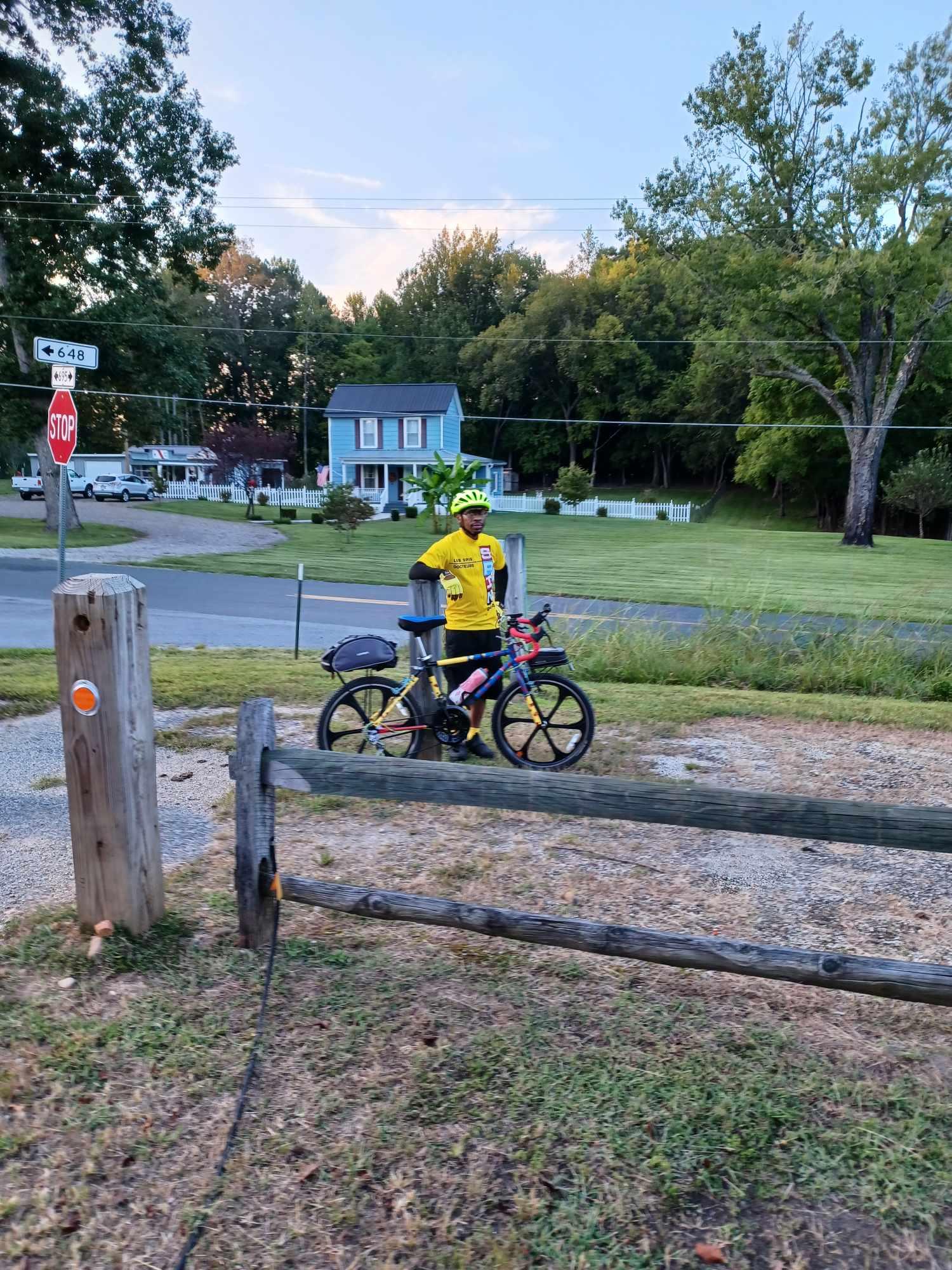 A person in a yellow shirt stands with a bicycle by a wooden fence.