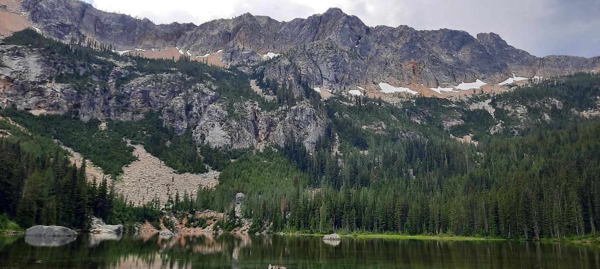 Calm mountain lake reflecting dense forest and rocky peaks under an overcast sky.