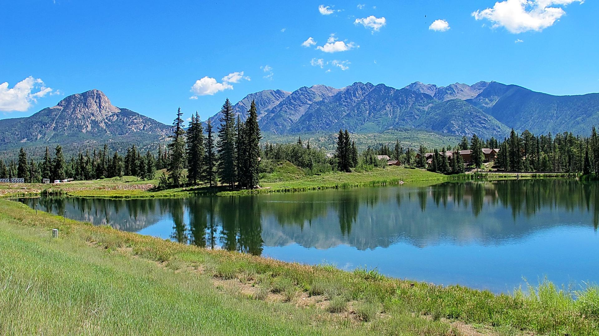Sunny day with a calm lake reflecting mountains and pine trees.