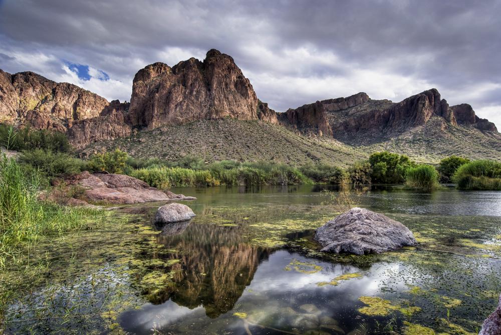 A tranquil lake with large rocks reflects dramatic mountains under a cloudy sky.