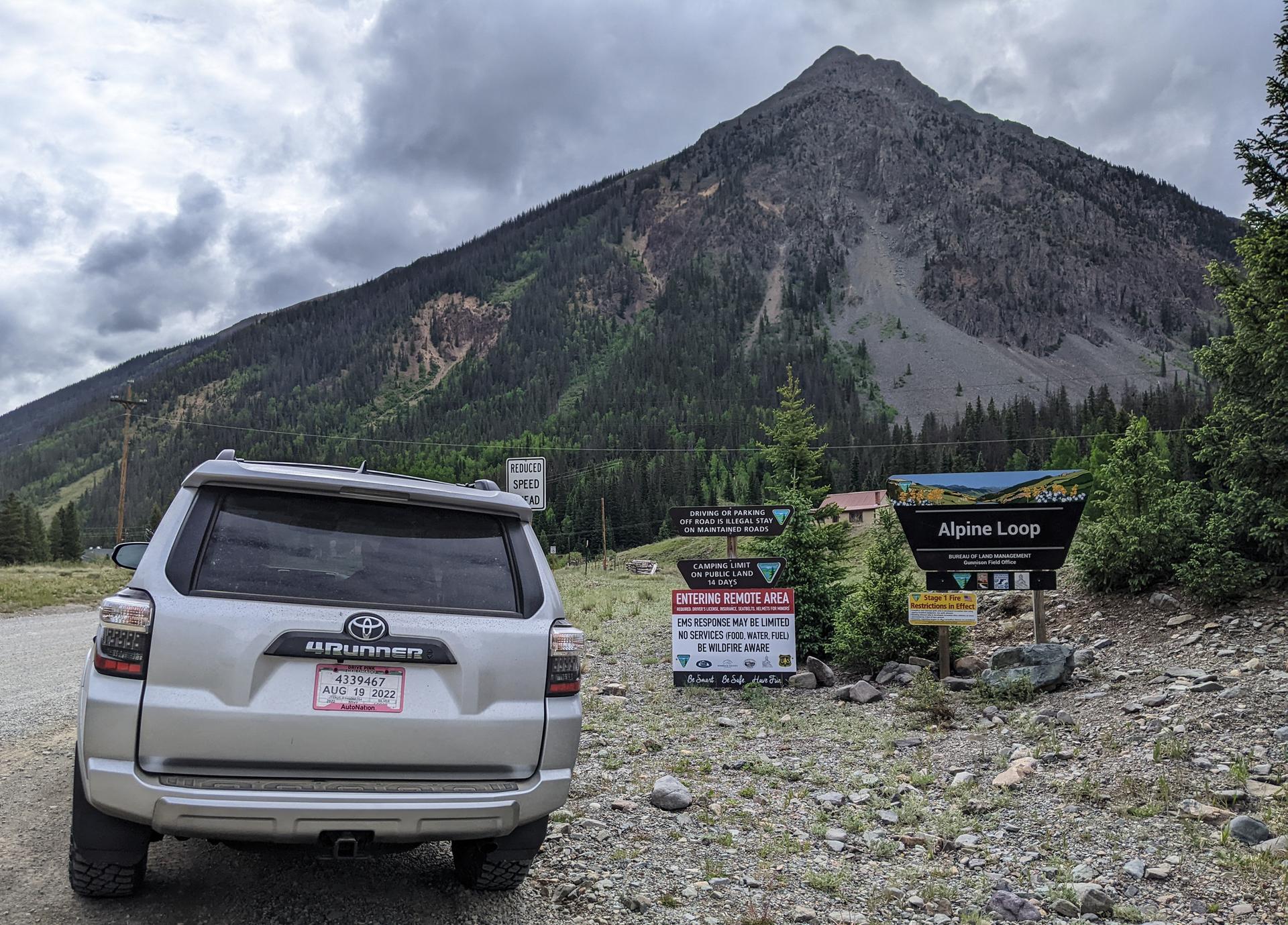 Silver SUV parked on dirt road near mountain under cloudy sky.