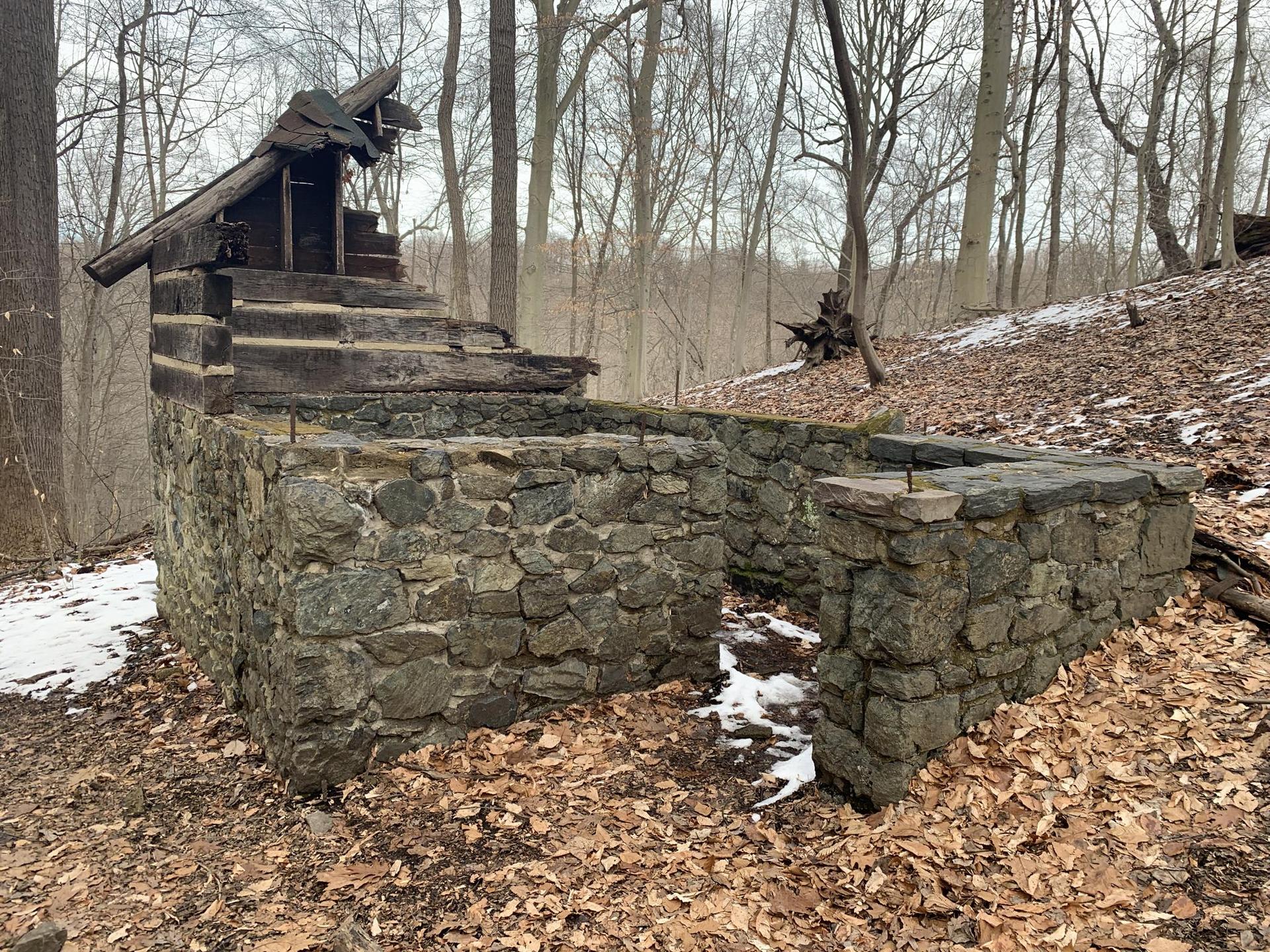 Stone and log ruins stand in a bare winter forest with patches of snow.