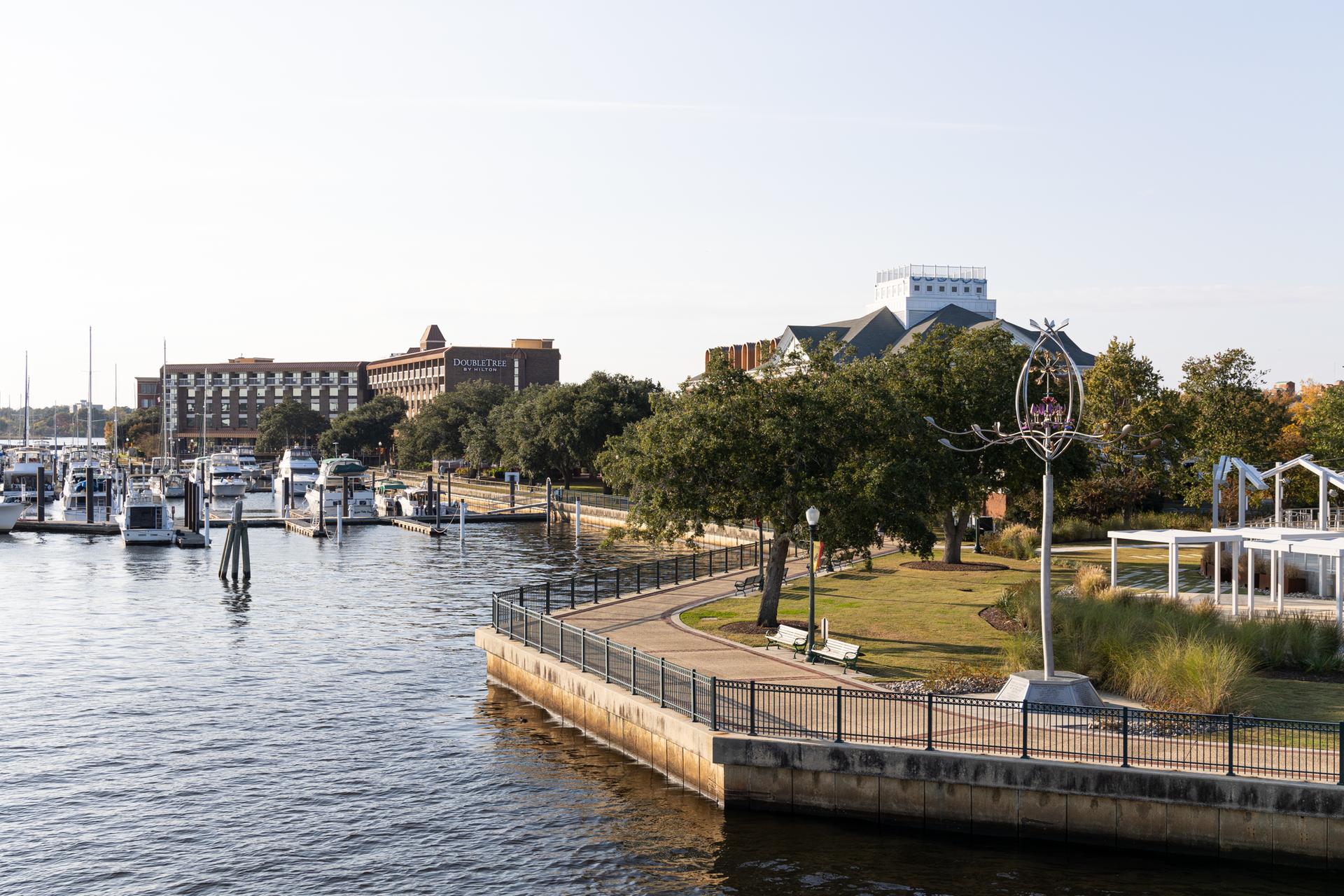 Marina with boats, waterfront path, green park, and buildings under bright sky.