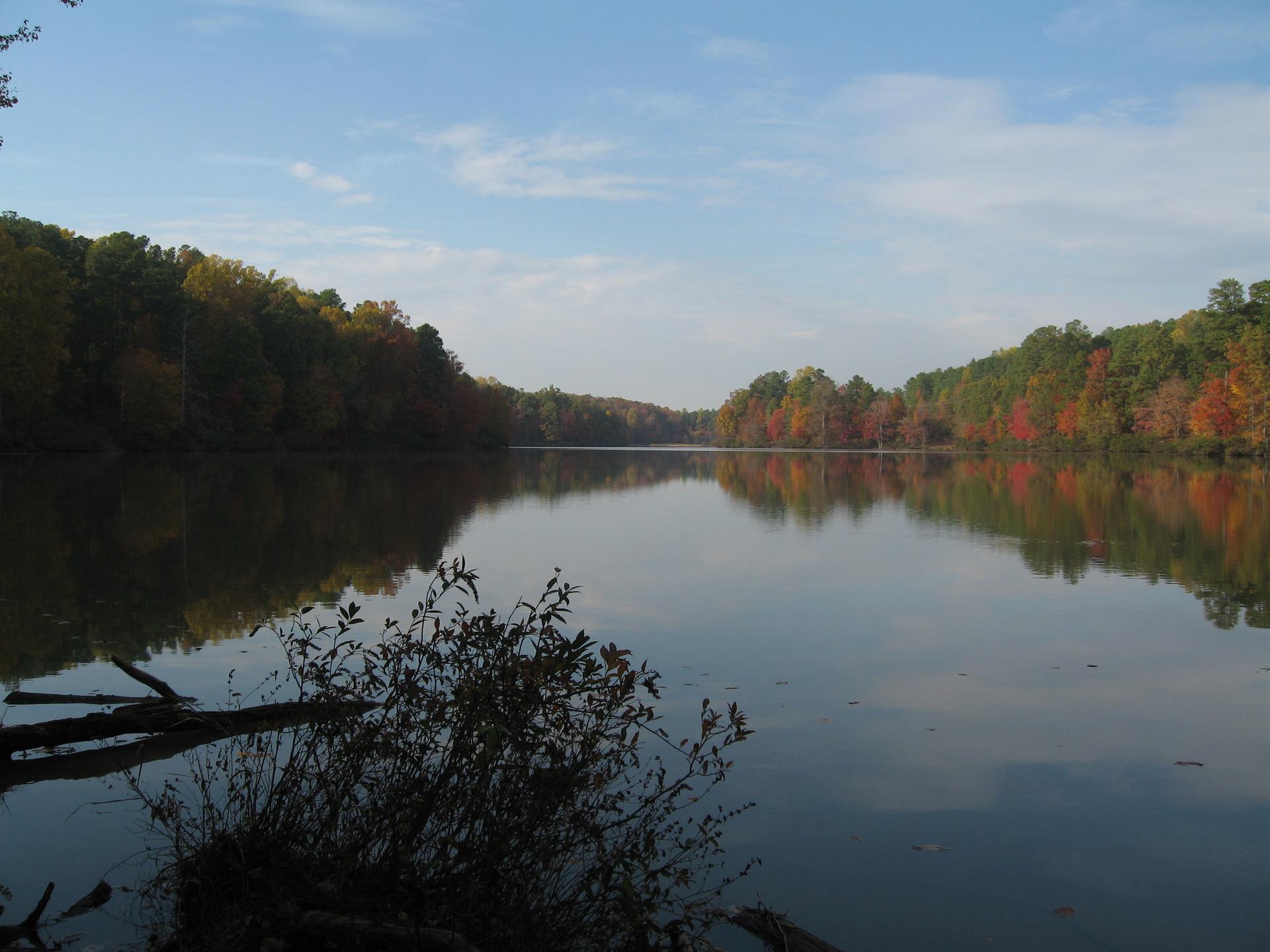 A wide, calm lake reflects colorful autumn trees under a blue sky.
