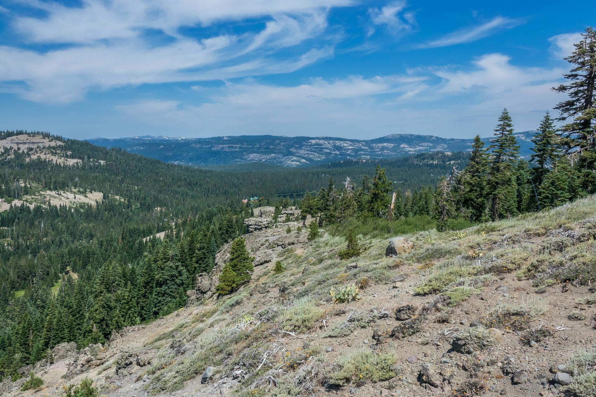 Wide mountain vista with forested slopes, rocky terrain, and a blue sky.
