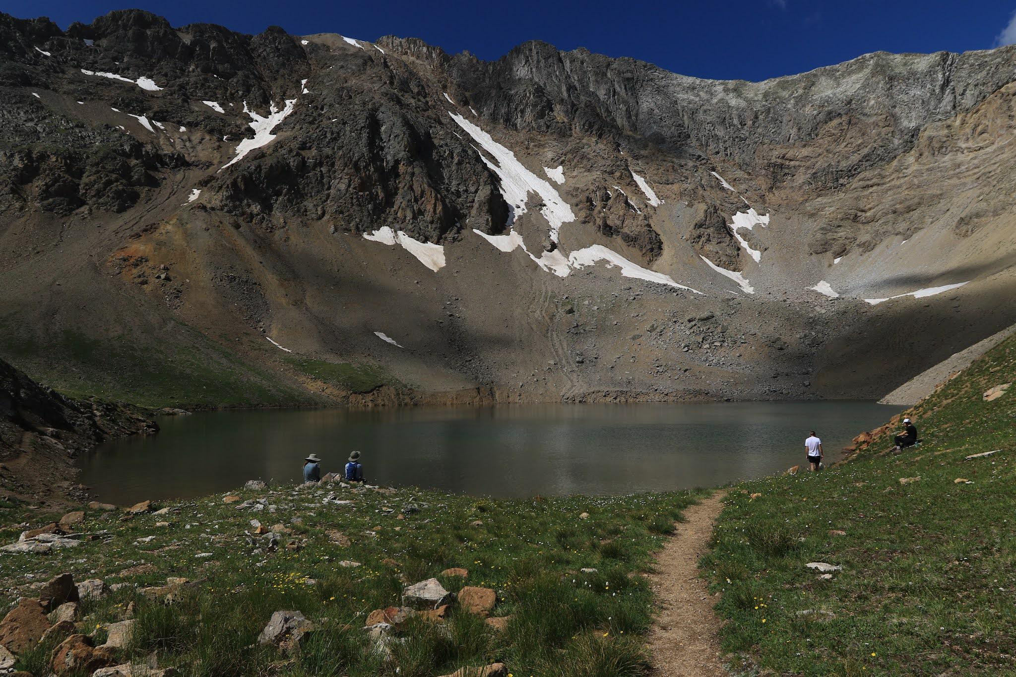 People relaxing by an alpine lake surrounded by rugged, snow-dusted mountains on a clear day.