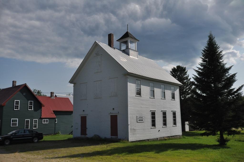 White building with cupola, green houses, and evergreen tree under cloudy sky.