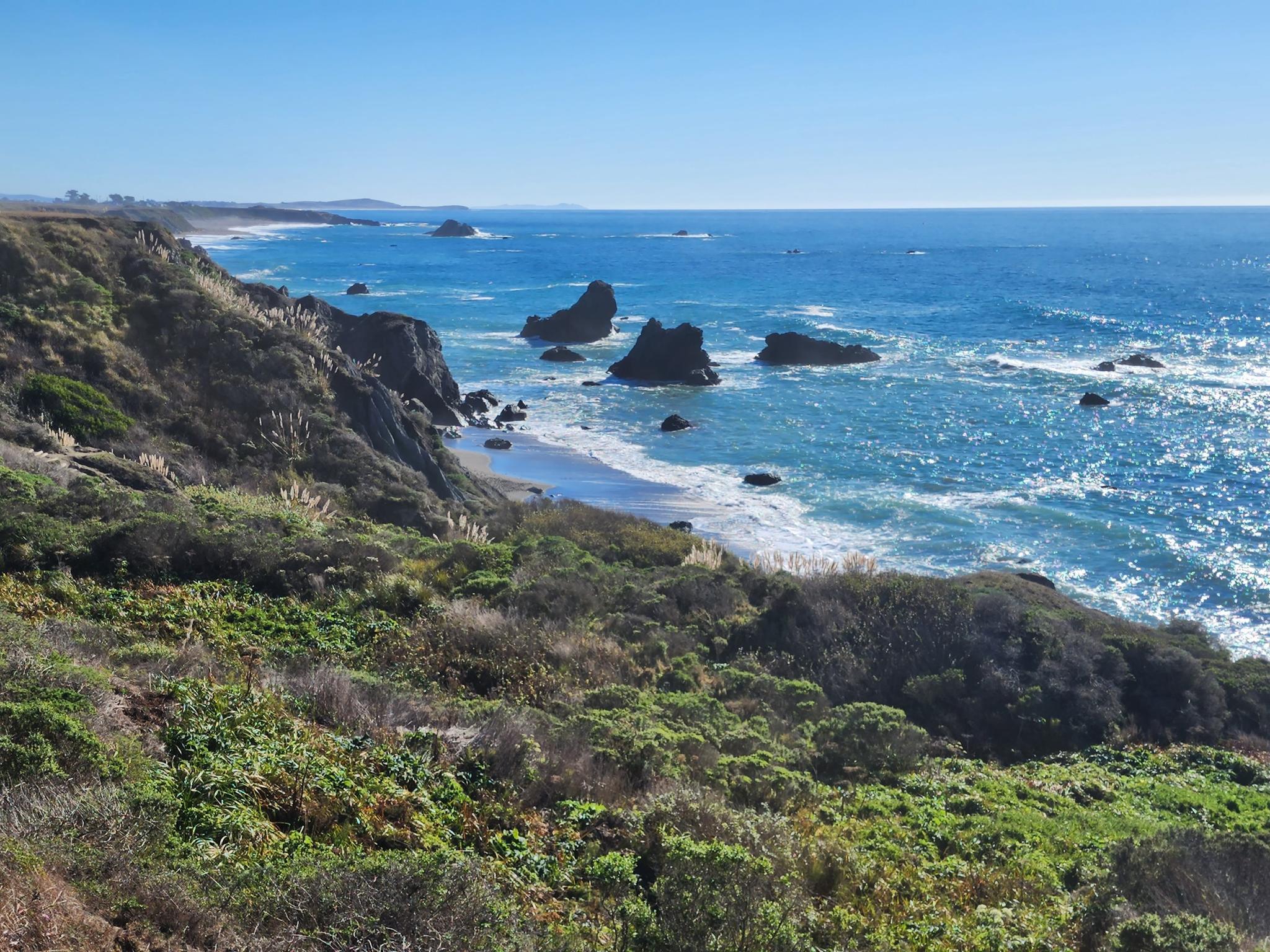 Bright coastal view with blue ocean, white waves, and green rocky cliffs.